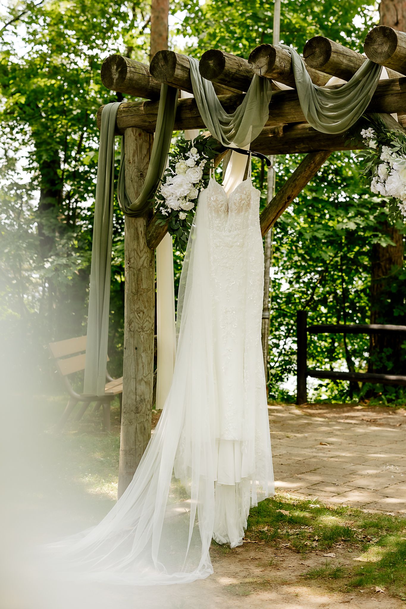 White lace wedding dress hanging on rustic wooden arch decorated with sage green draping and white flowers outdoors.