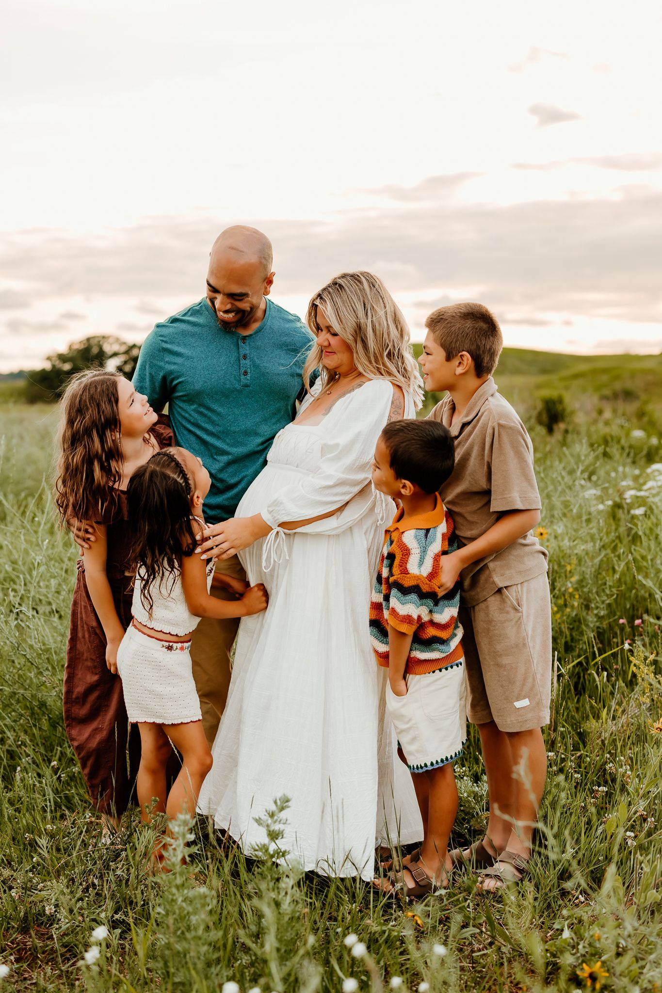A pregnant woman in a white dress stands with her husband and four children in a green field at golden hour sunset.
