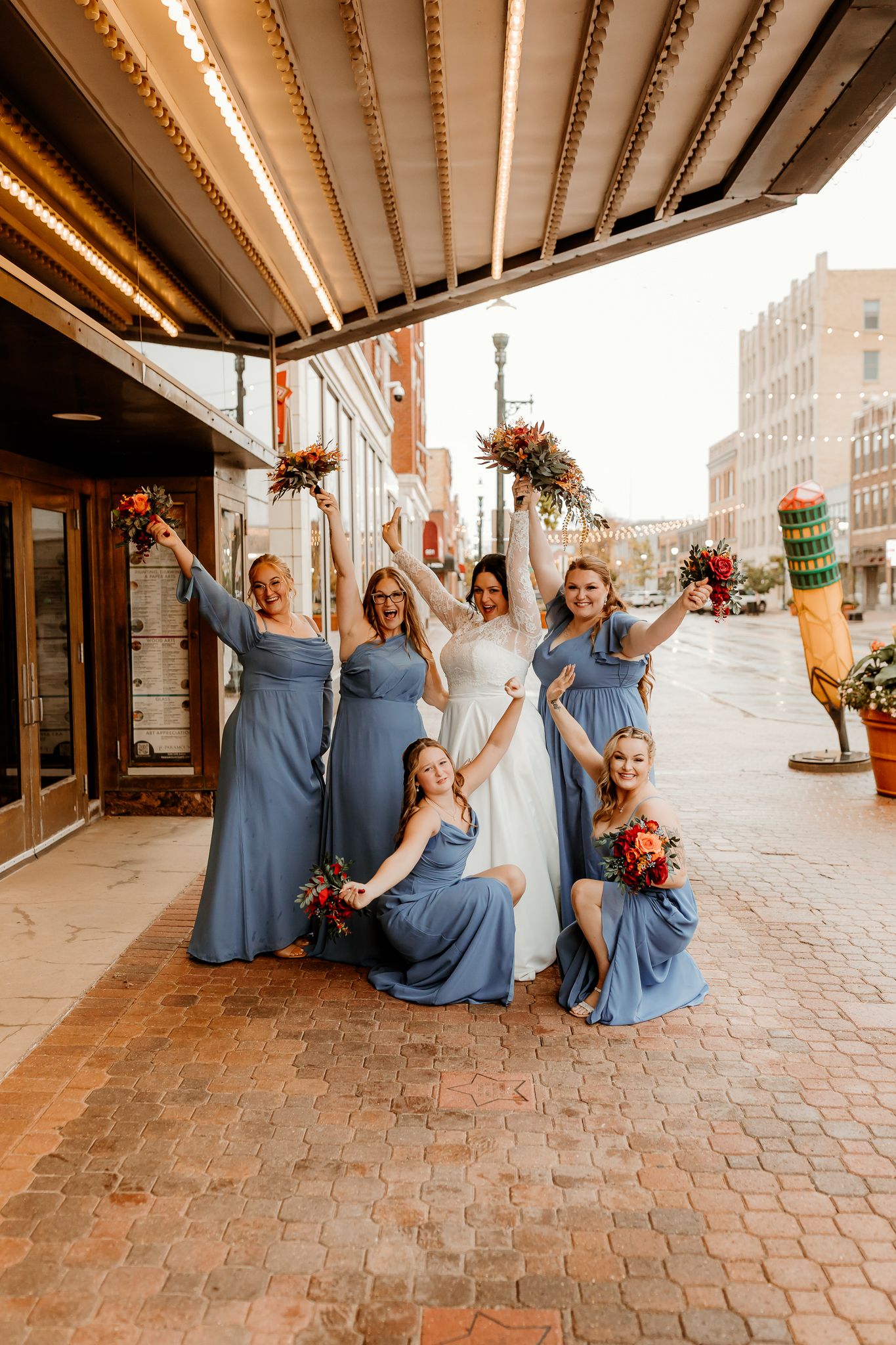 Bride in white gown celebrates with four bridesmaids in dusty blue dresses holding red bouquets on a brick street outside a theater.
