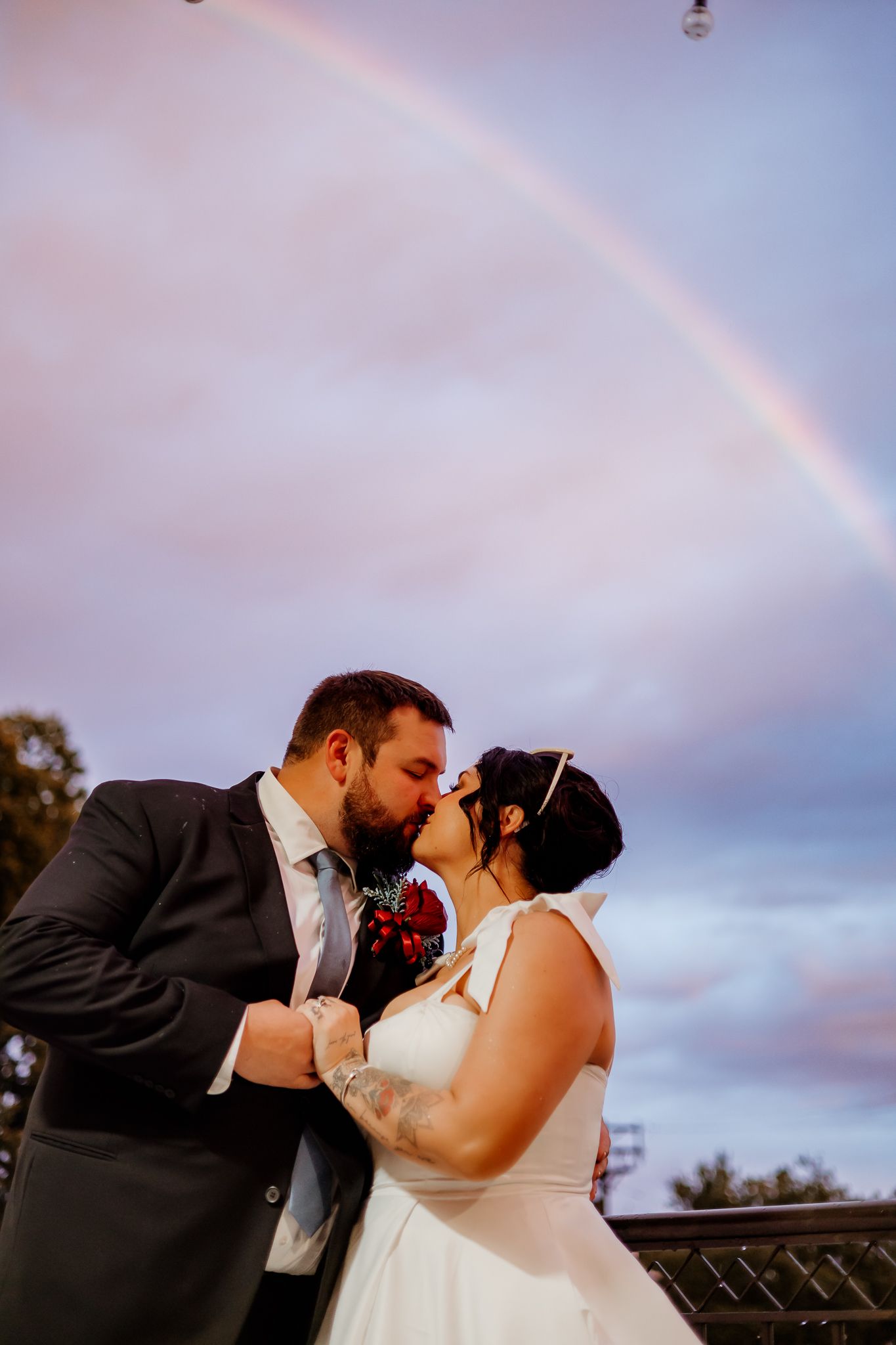 Bride in a white dress and groom in a gray suit share a kiss at sunset beneath a faint rainbow arching across the purple sky.