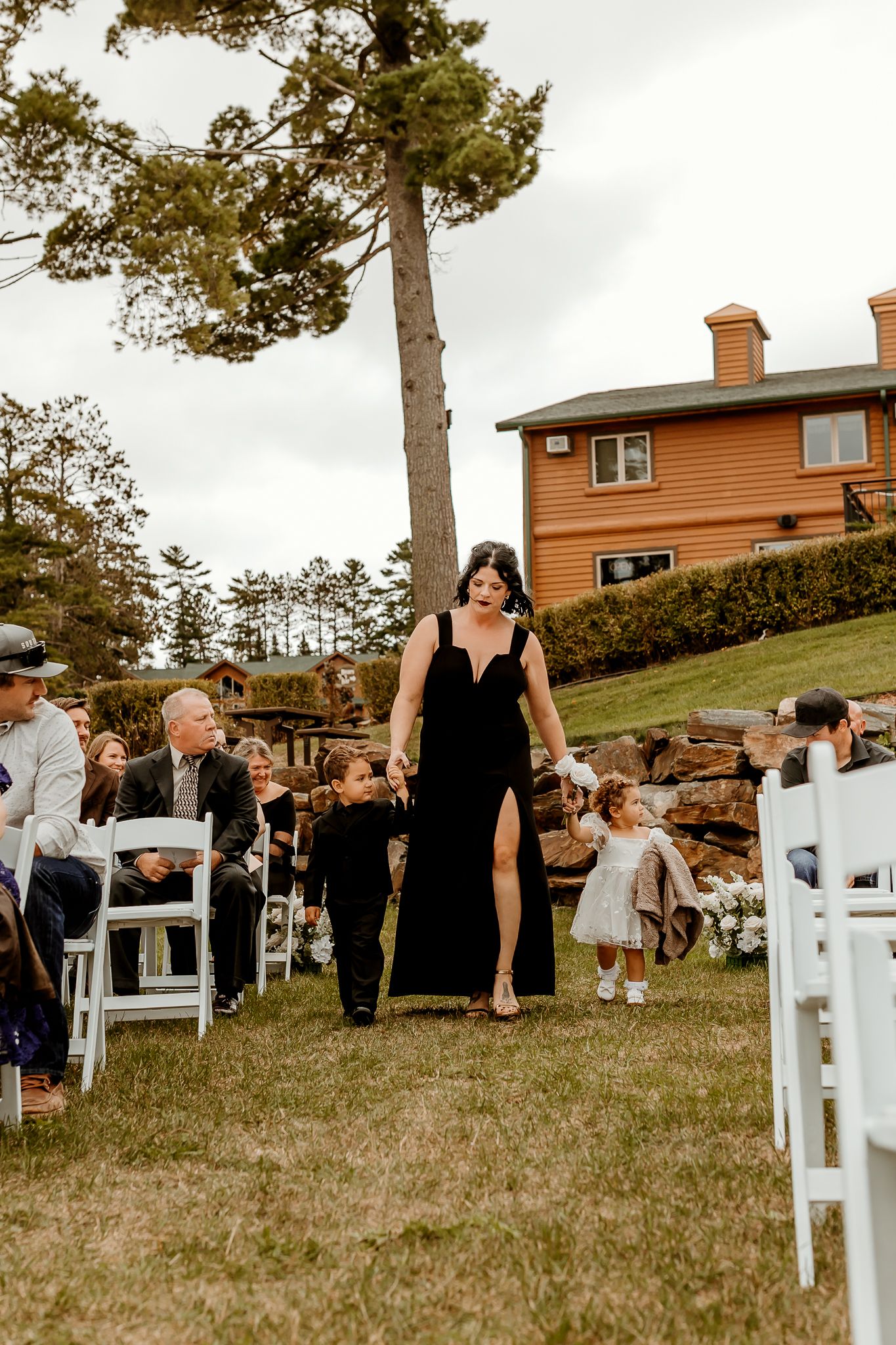 A woman in a black dress walks two toddlers down a grassy outdoor wedding aisle lined with white chairs and seated guests.