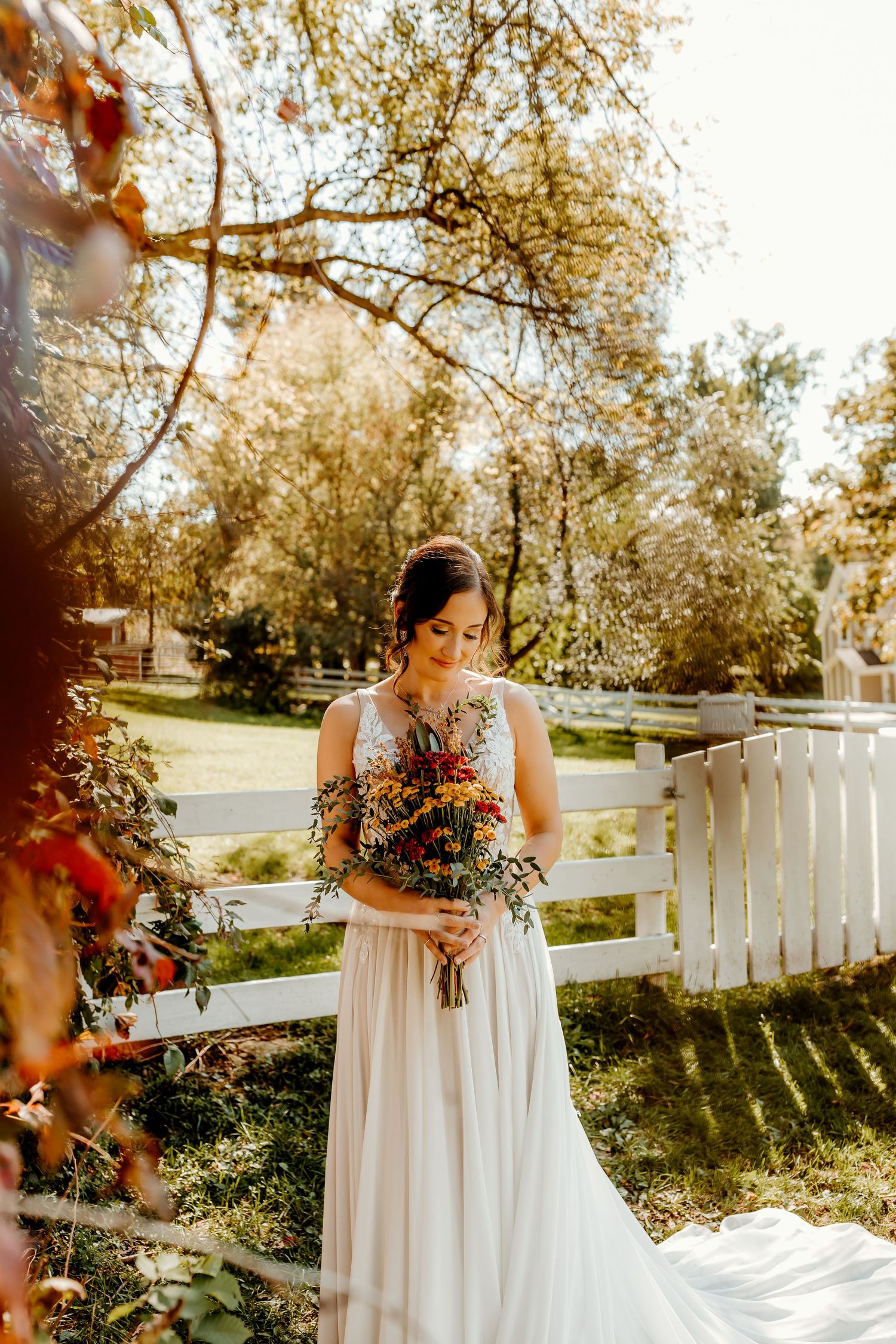 Bride in a flowing white gown holds a colorful autumn bouquet while standing by a white picket fence in a sunlit garden.