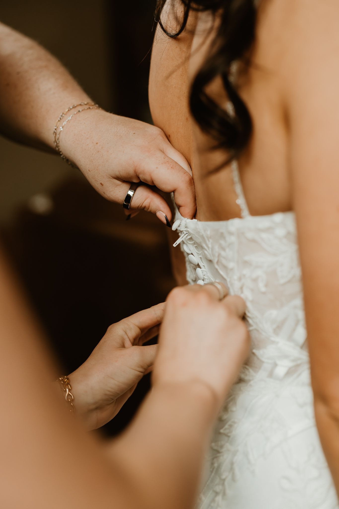 Two women's hands fasten the lace-up back of a white floral lace wedding gown on a bride with long dark hair.