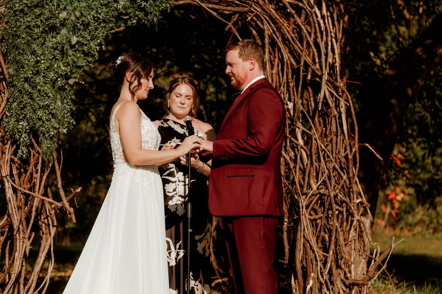 Bride in a white lace gown and groom in a burgundy suit exchange vows with an officiant under a rustic woven branch wedding arch.