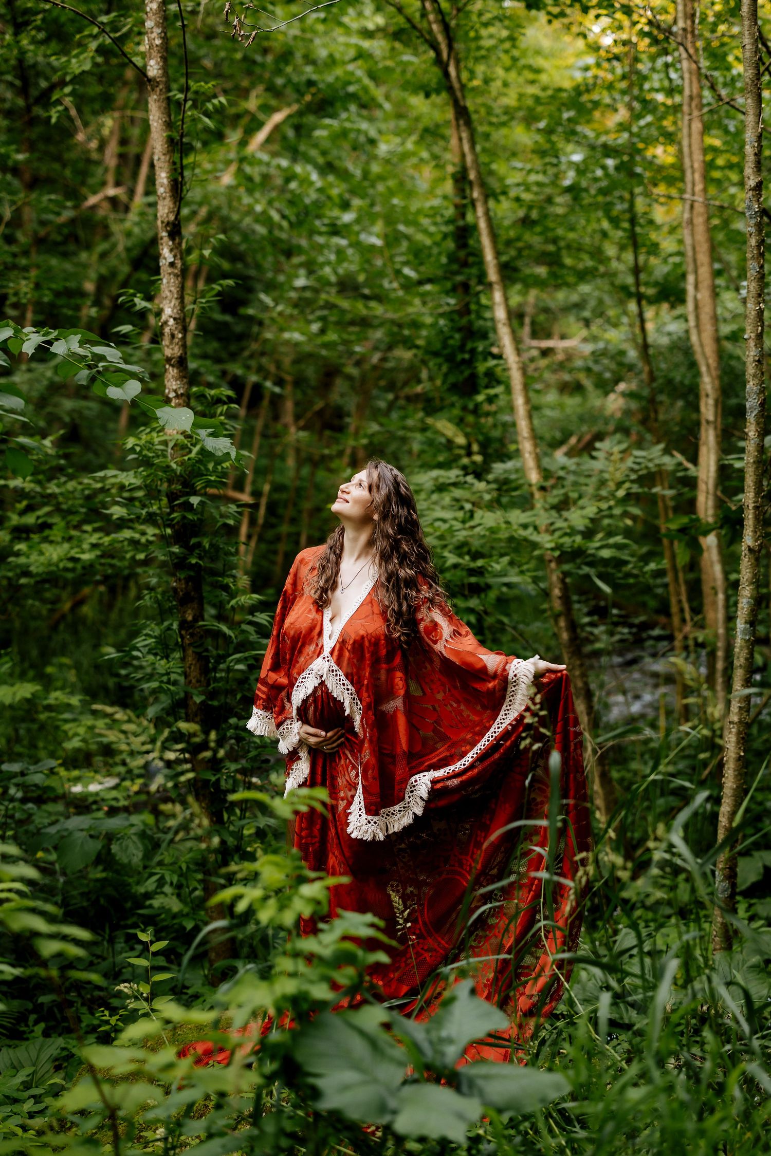 Woman in flowing red boho dress standing in lush green forest, looking up with arms outstretched.