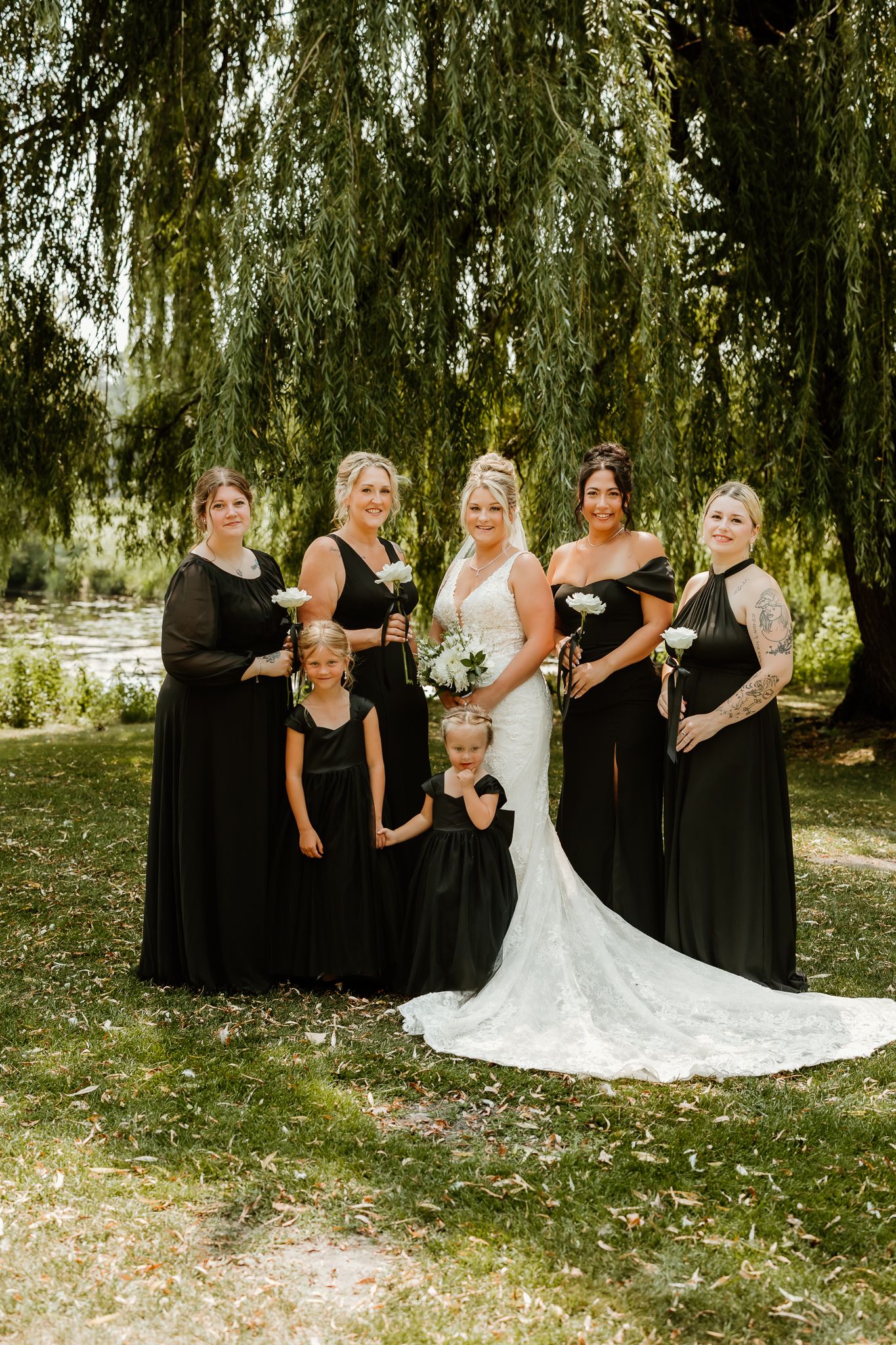 Bride in white gown poses with bridesmaids in black dresses and flower girls under a weeping willow tree.