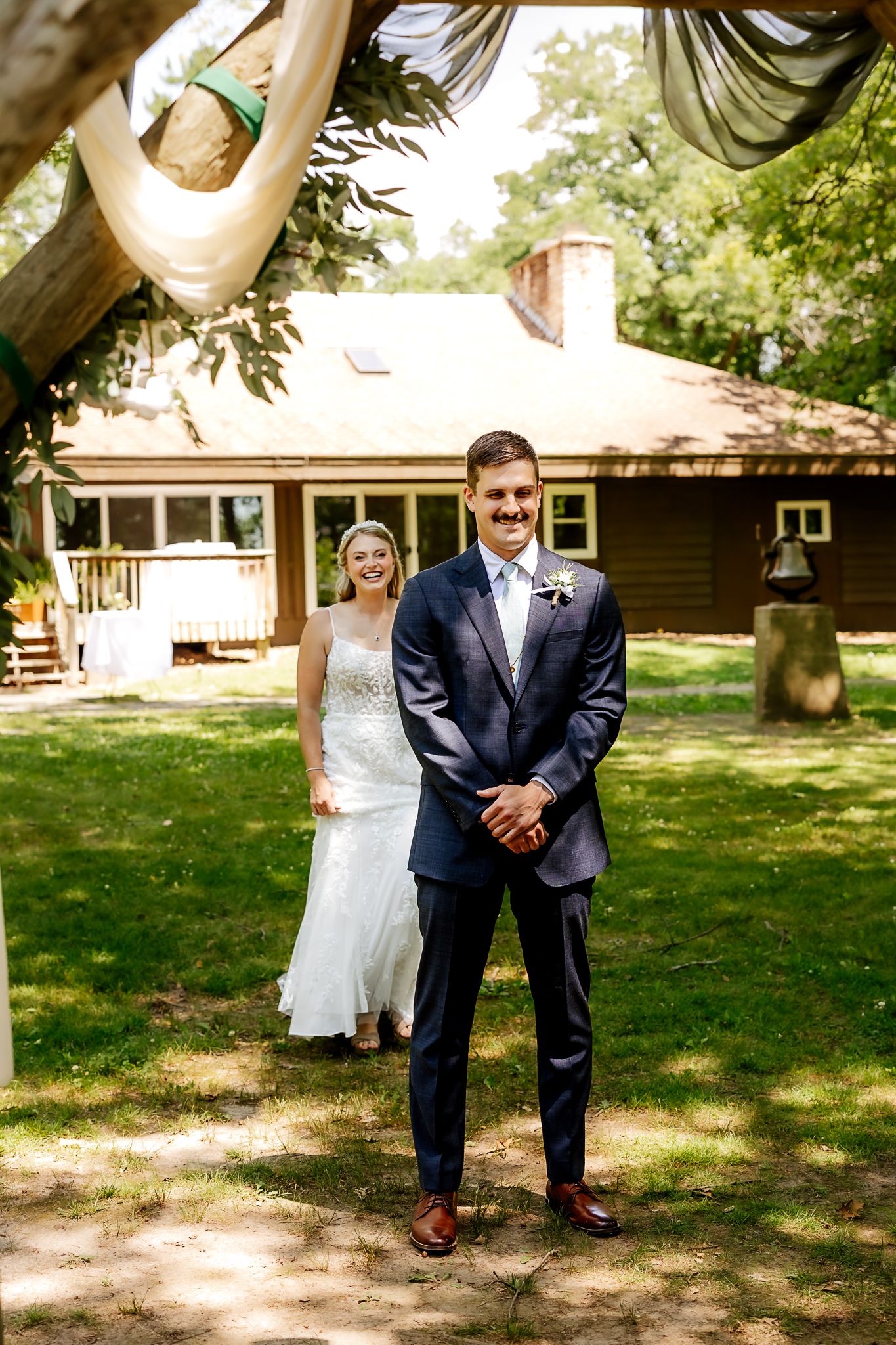 Bride in white gown and groom in navy suit share a first look moment outdoors near a rustic cabin.