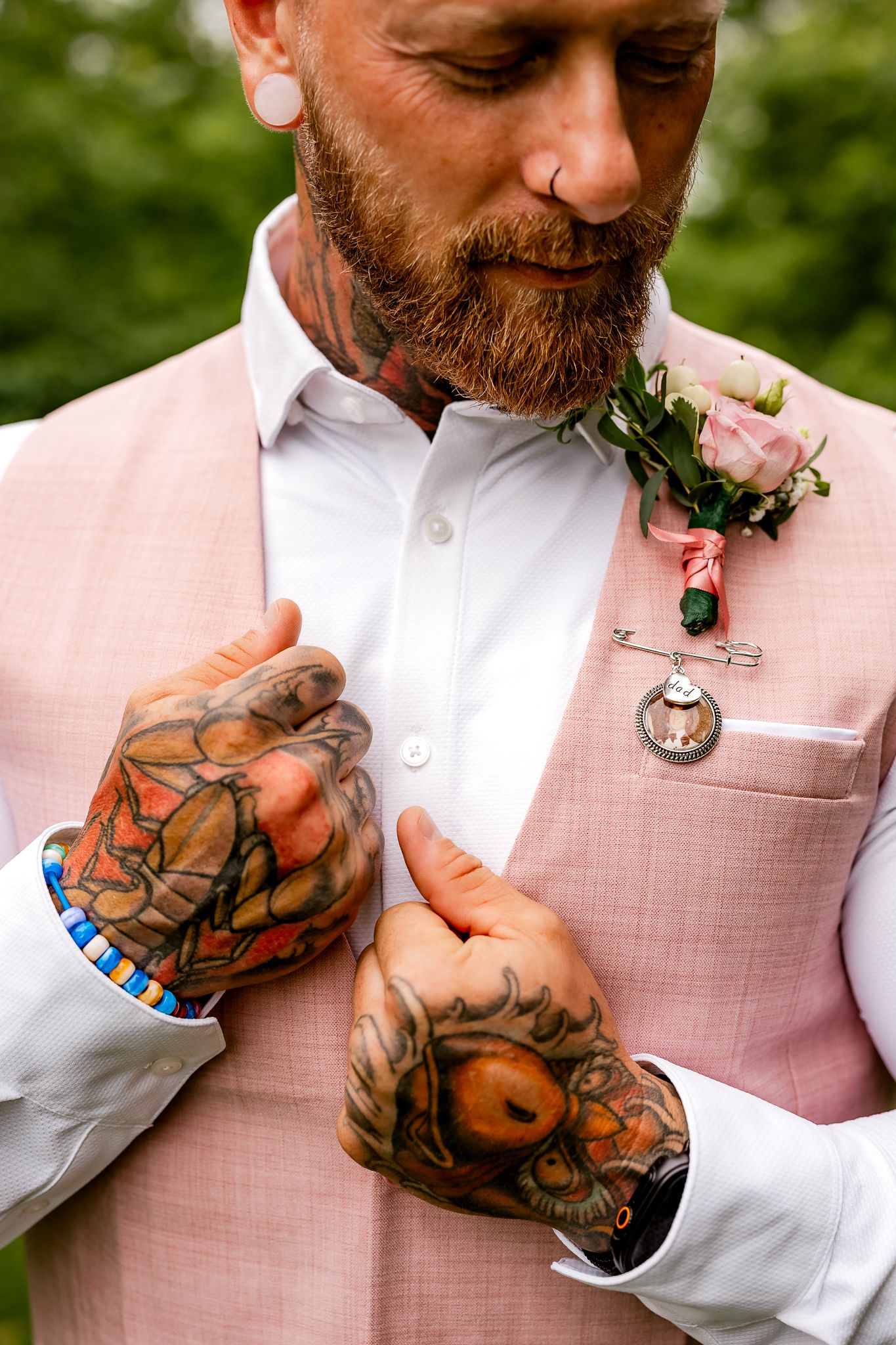 Tattooed groom in pink suit adjusting boutonniere with pocket watch pin at wedding ceremony.