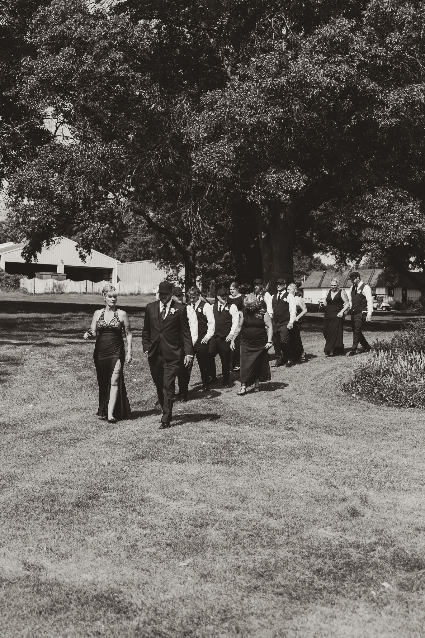 Black and white photo of a wedding party walking across a grassy lawn beneath large trees.