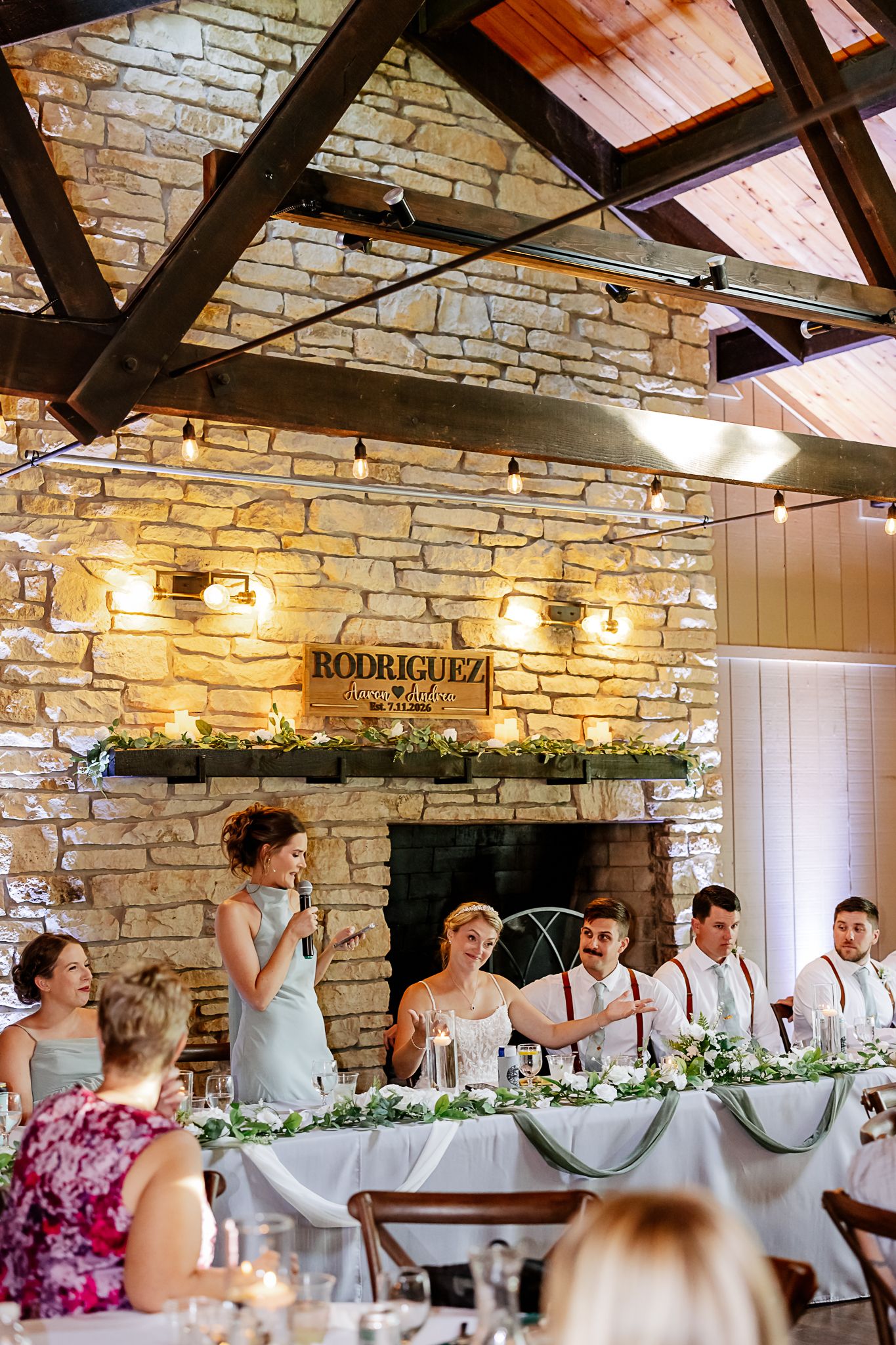 Wedding reception at rustic stone venue with string lights, bridal party at head table, and Rodriguez wedding sign above fireplace.
