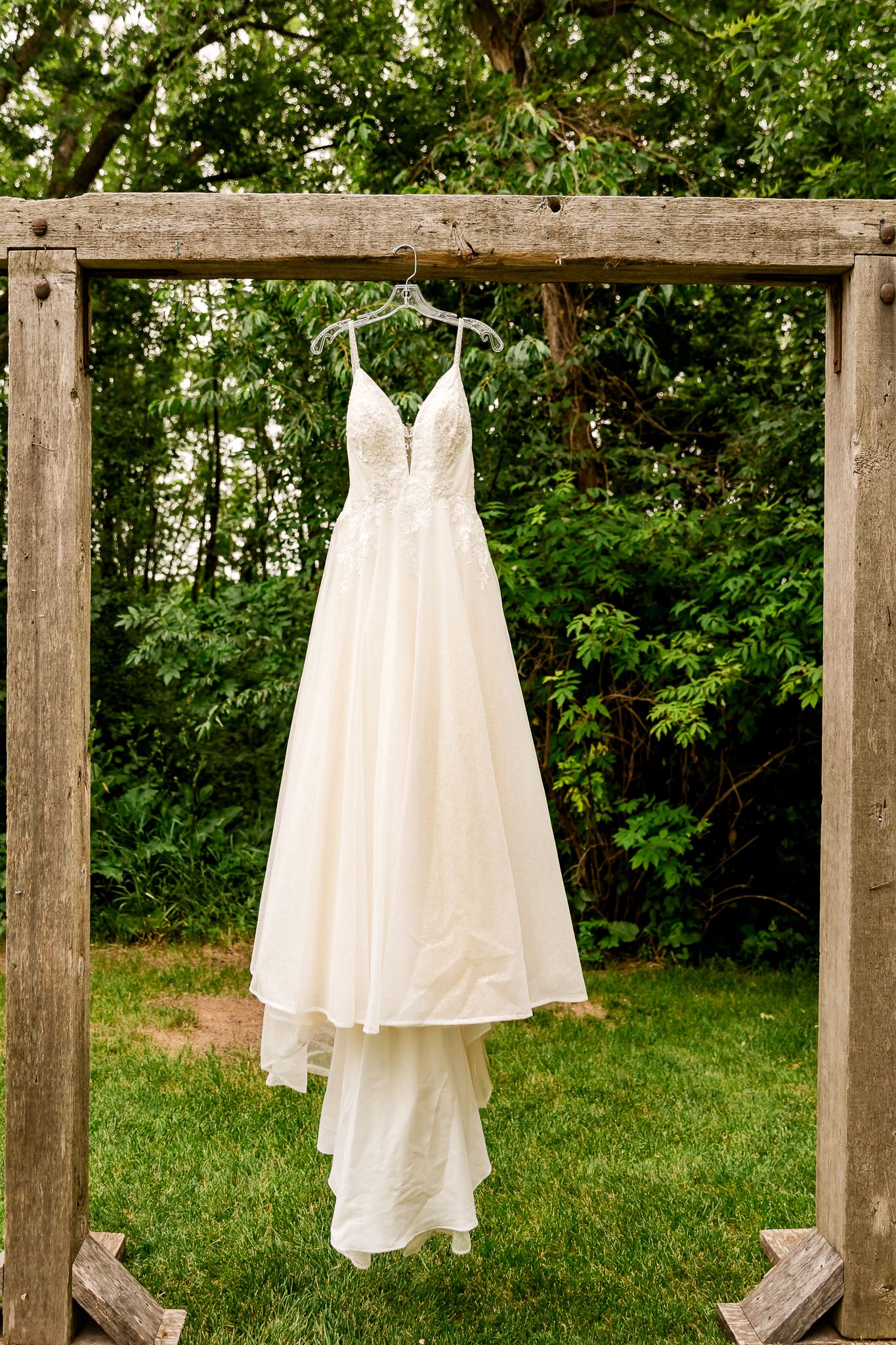 Elegant ivory wedding dress with lace bodice hanging on wooden arch frame surrounded by lush green trees outdoors.