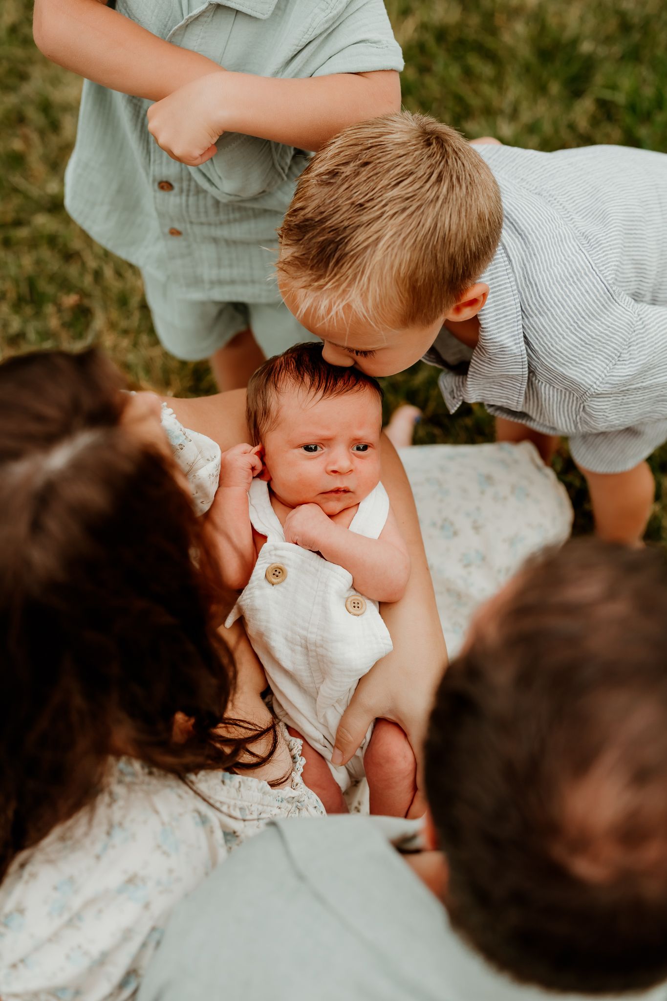 Family gathered around newborn baby as older sibling leans in to give a gentle kiss outdoors.