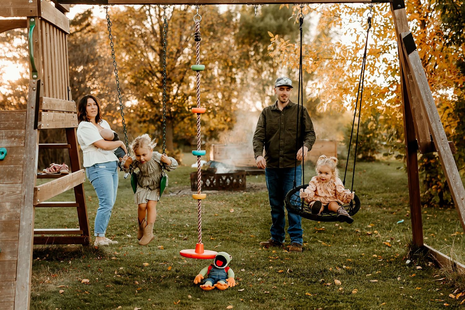 Family of four enjoying a backyard playset in autumn, with a toddler on a swing, a child playing, and a bonfire glowing in the background.