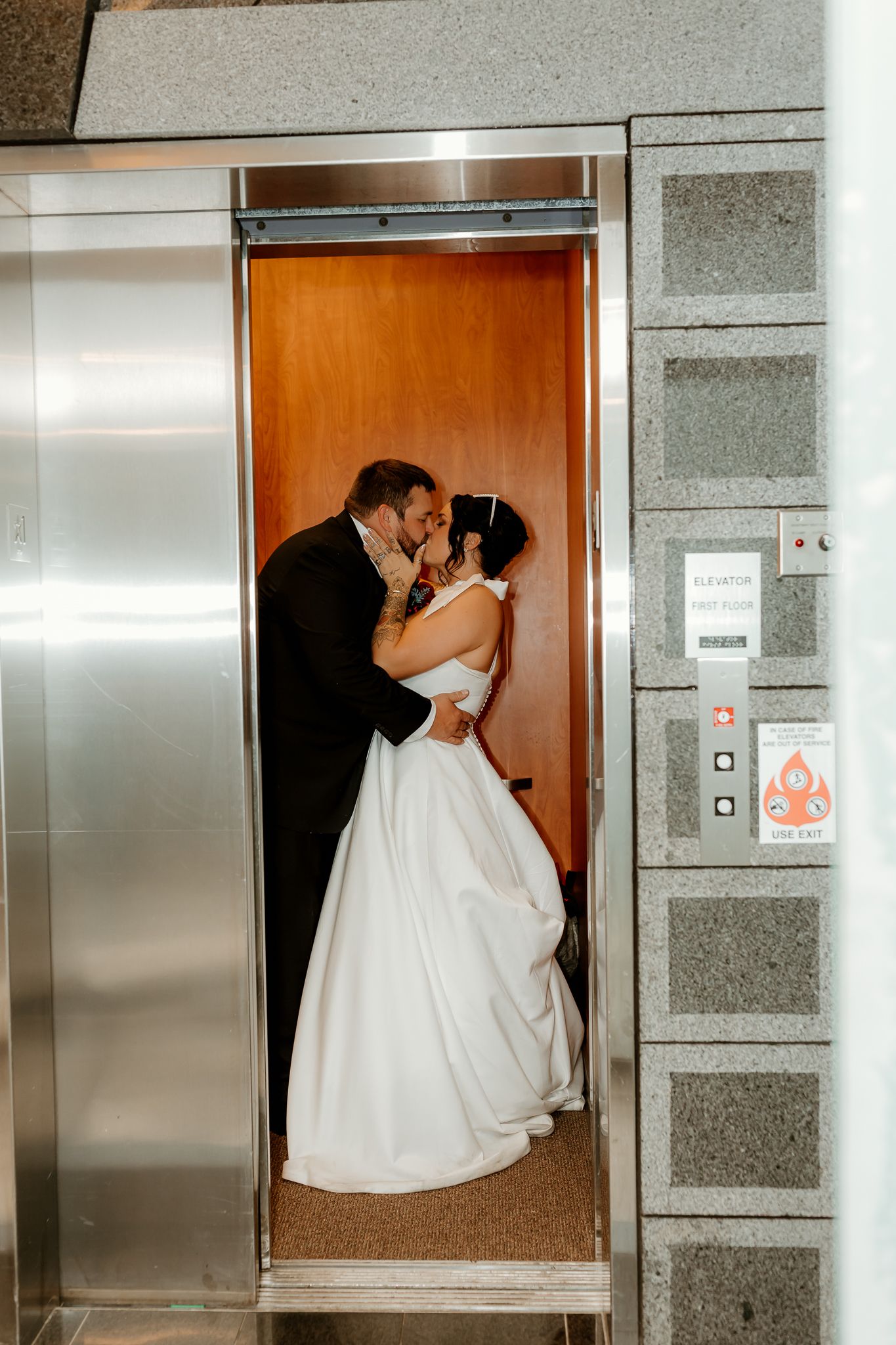 Bride in a white gown and groom in a black suit share a kiss inside an open wood-paneled elevator on their wedding day.