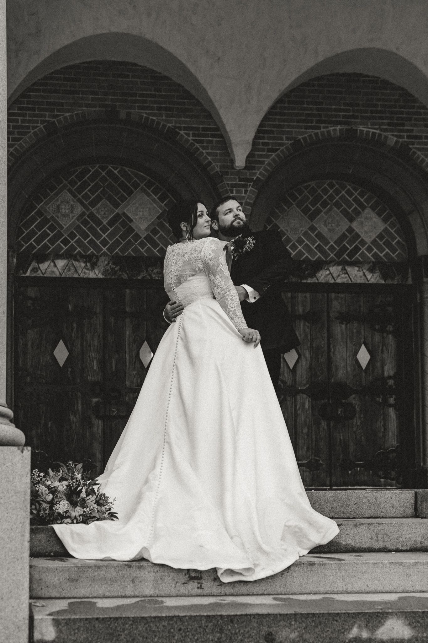 Black and white wedding photo of a bride in a lace-sleeved gown embracing her groom on stone church steps before ornate arched wooden doors.