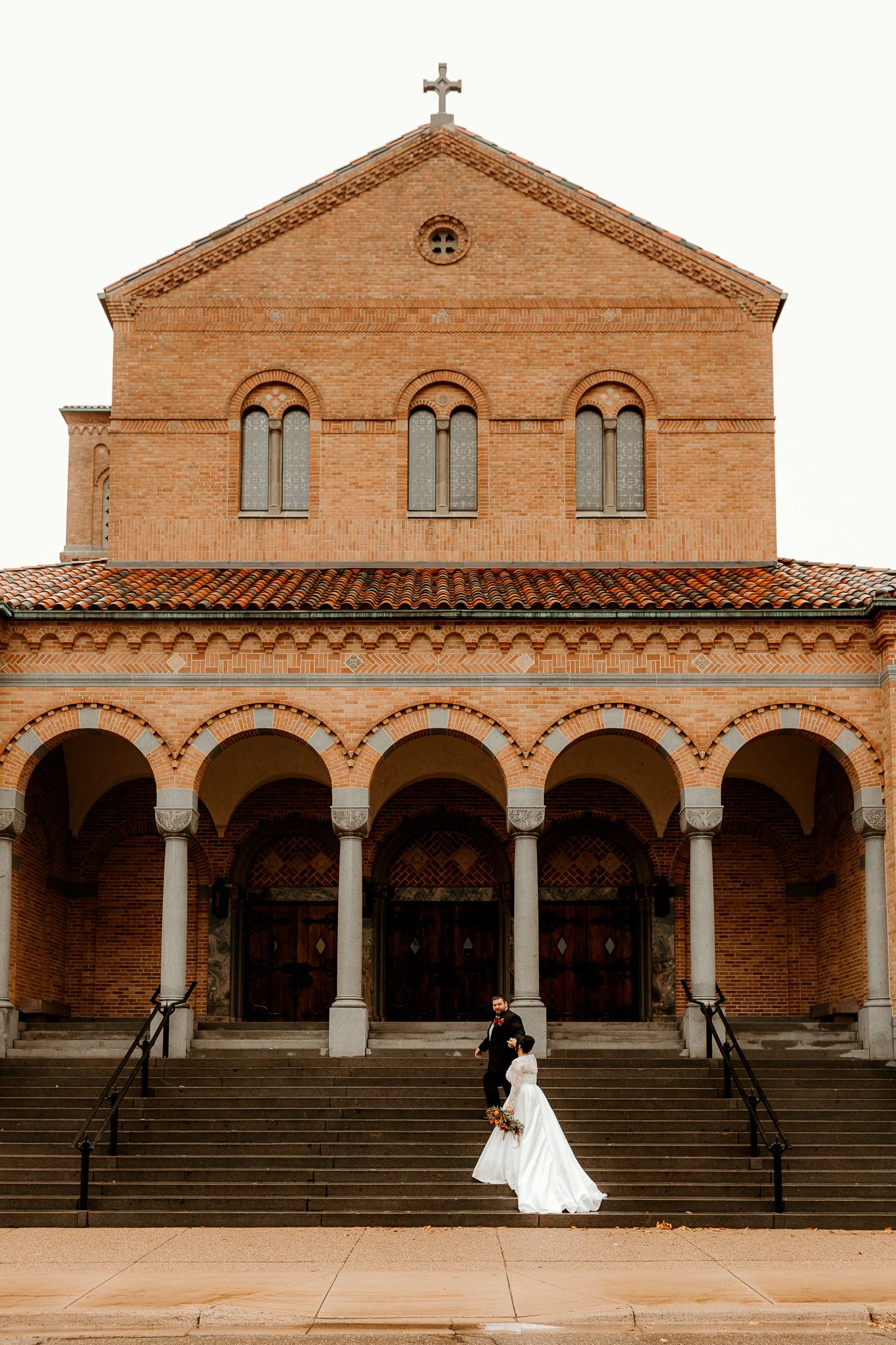 Bride in a white gown and groom in a dark suit stand on the steps of a brick church with arched entrances and a cross on top.