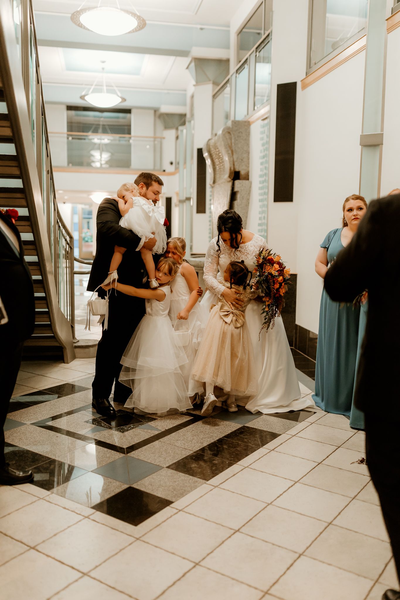 Bride in a white gown and groom in a dark suit embrace two flower girls in a grand lobby with a checkered marble floor.