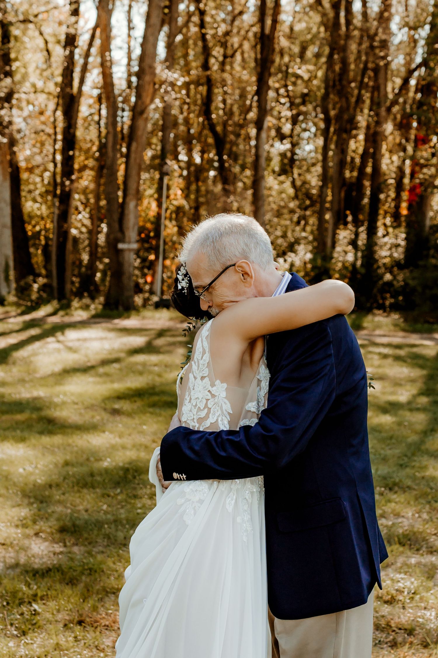Elderly man in a navy blazer embraces a bride in a lace wedding gown in a sunlit wooded outdoor setting.