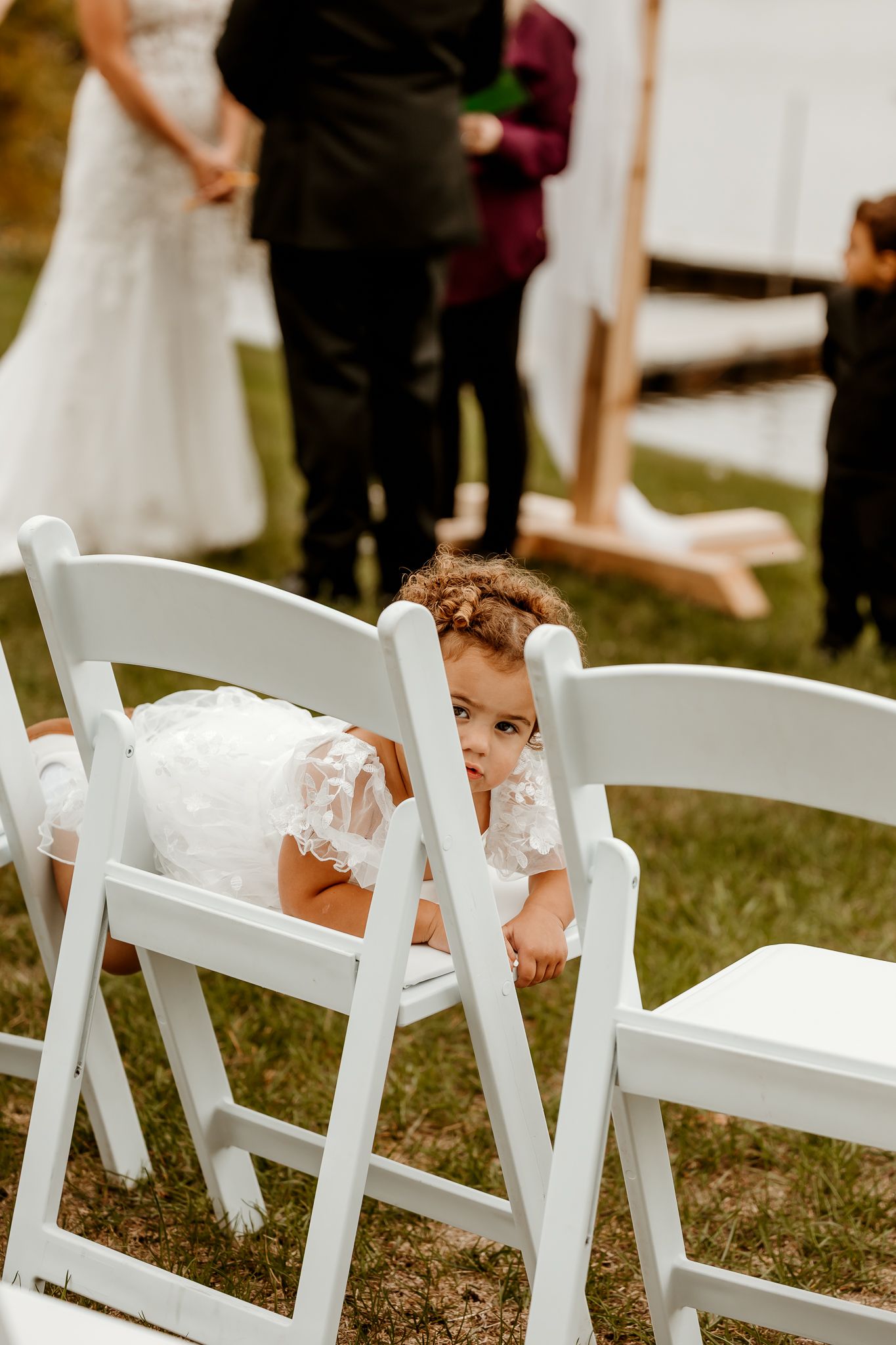 Toddler in a white lace dress peeks over a white folding chair at an outdoor wedding ceremony while the bride and guests stand nearby.