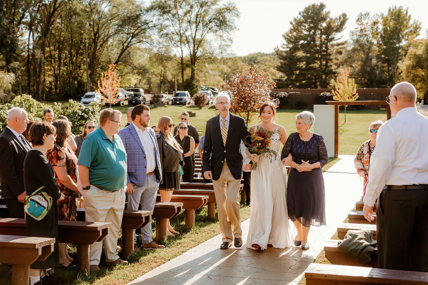 Bride in a white gown and groom in a gray suit walk down an outdoor wedding aisle past seated guests on wooden benches.