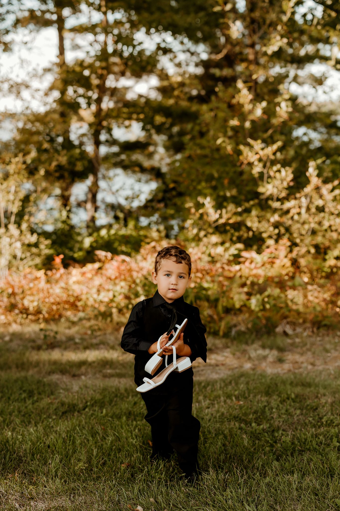 Young boy in a black button-up shirt and pants holds a pair of white sandals while standing on grass near colorful autumn foliage.