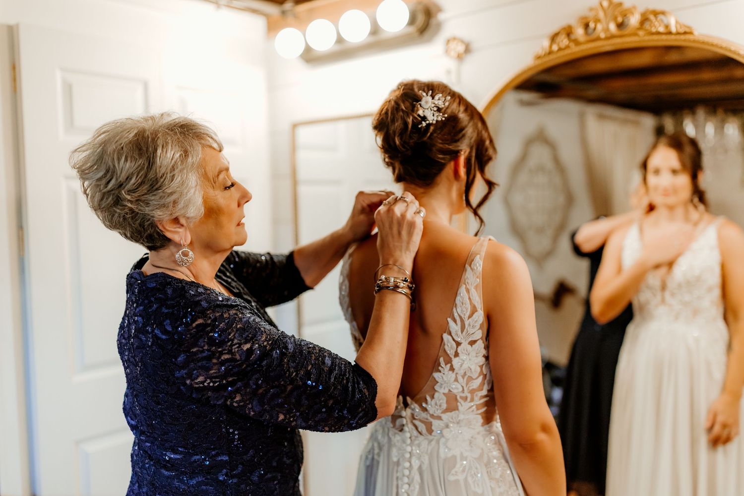 Mother in a navy lace dress helps a bride in a beaded lace wedding gown with her jewelry as another woman looks on.