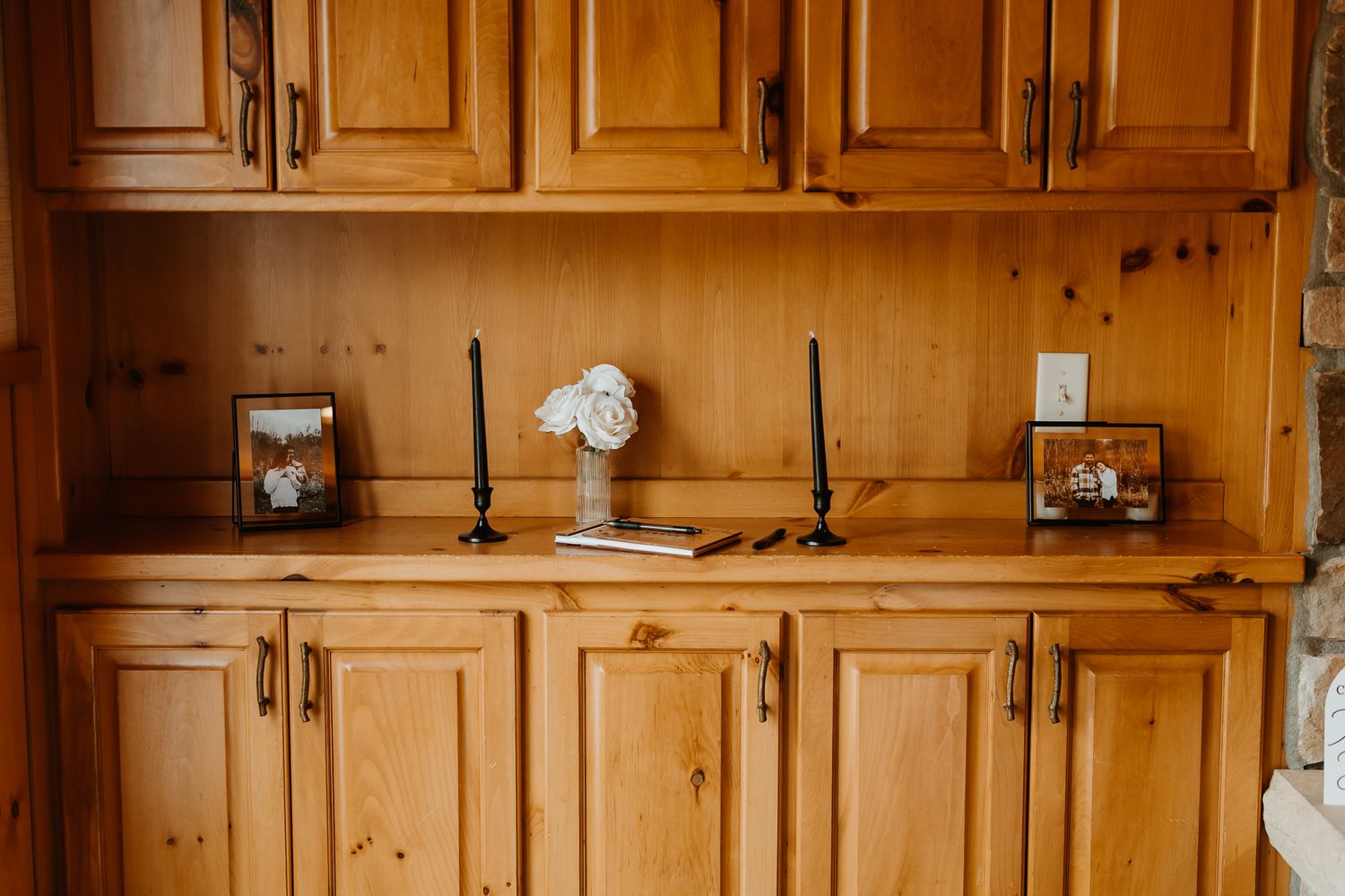 Rustic knotty pine built-in cabinet with black candlesticks, a white rose in a vase, and two framed shadow boxes on the wooden countertop.