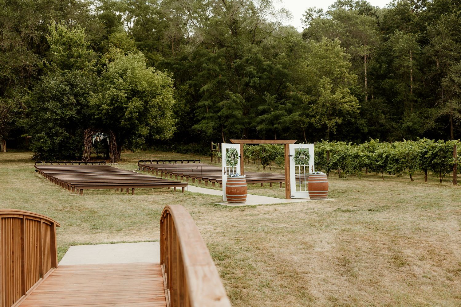 Outdoor vineyard wedding ceremony setup with wooden barrel arch, bench seating, and lush green trees in background.