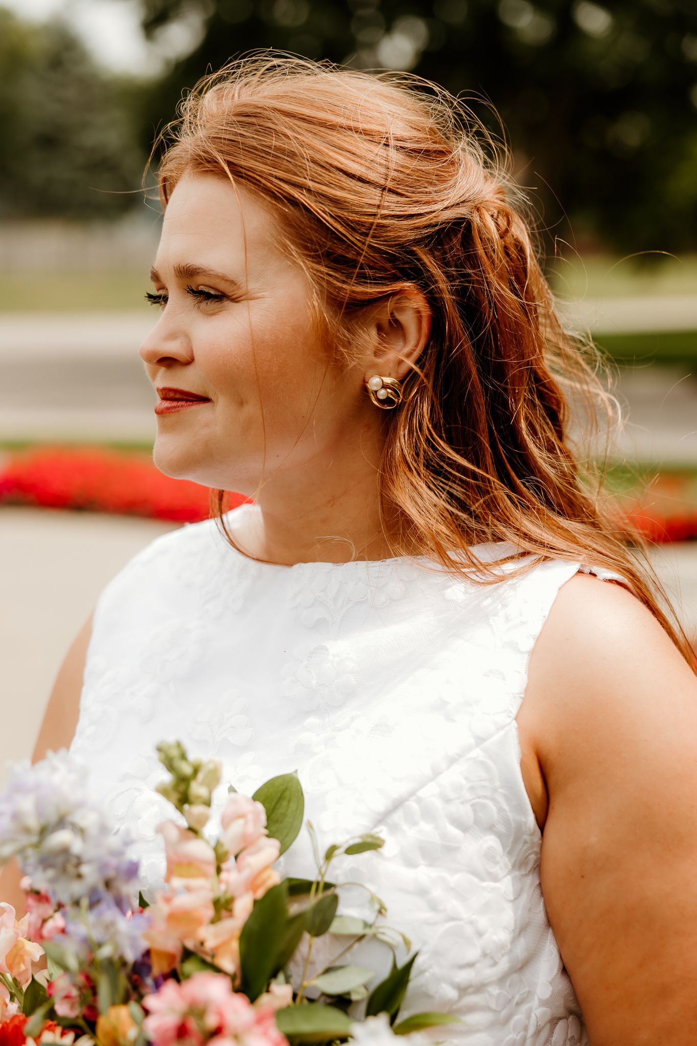 Bride with red hair in white dress holding colorful bouquet, wearing pearl earrings, smiling softly outdoors.