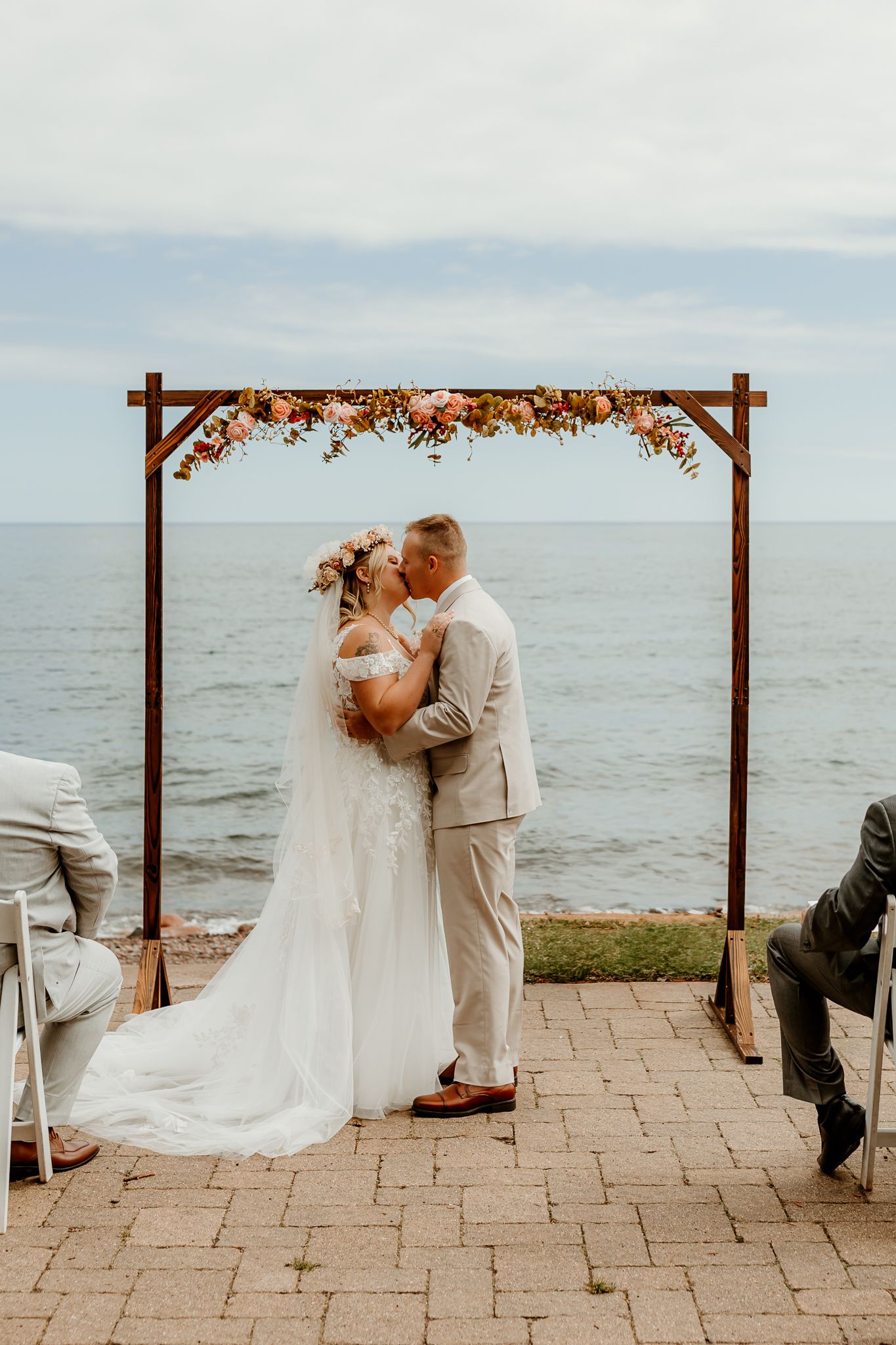 Bride and groom share first kiss under floral arch by the ocean at outdoor waterfront wedding ceremony.