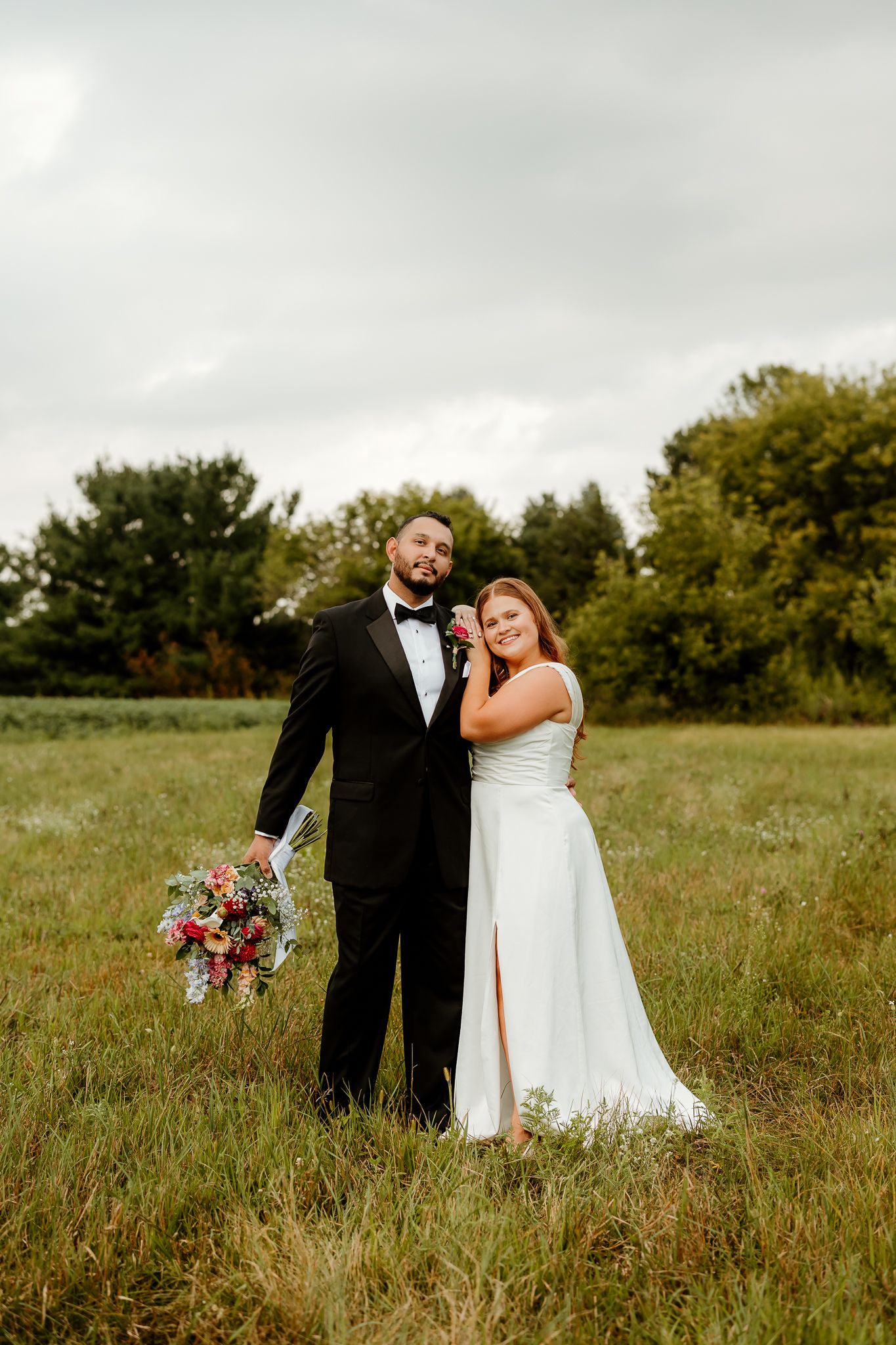 Bride and groom posing in a green field, groom in black tuxedo holding bouquet, bride in white gown on cloudy day.