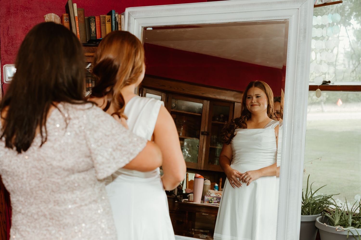 Bride in white gown views her reflection in a large mirror while a woman in lace dress stands beside her.