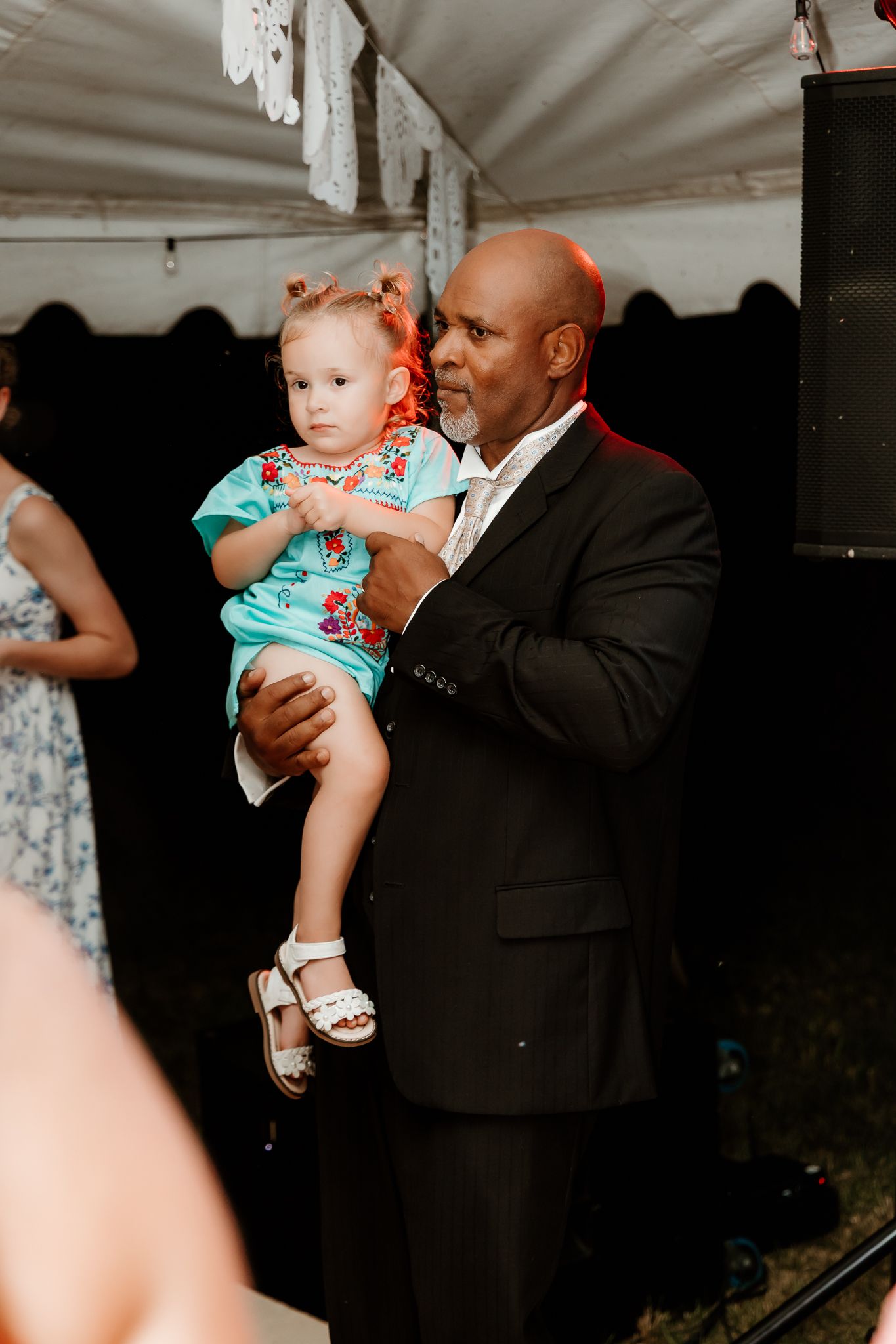 A man in a black suit holds a toddler girl in a floral dress at an outdoor event under a tent.