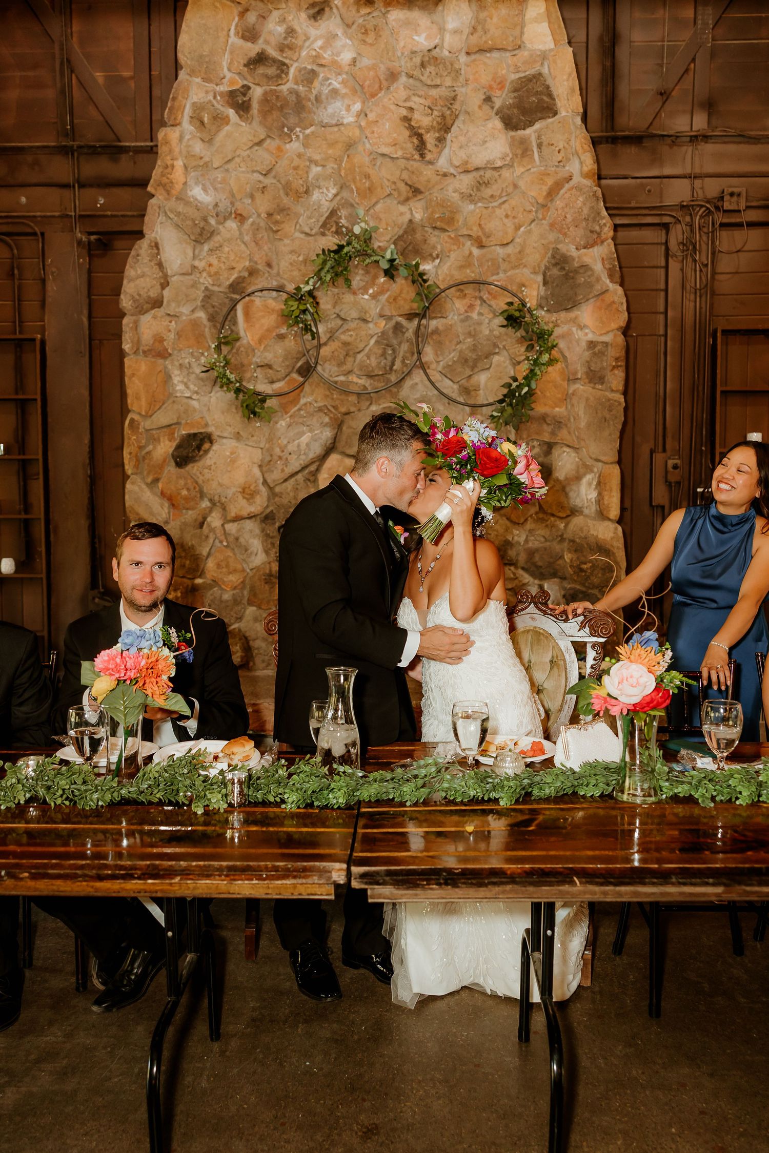 Bride and groom share a kiss at rustic wedding reception sweetheart table with colorful floral arrangements and stone fireplace backdrop.