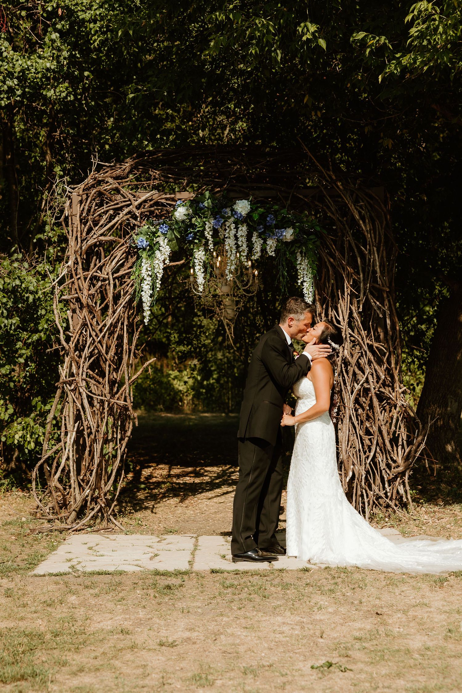 Bride and groom sharing first kiss under rustic twig arch adorned with wisteria flowers at outdoor wedding ceremony.