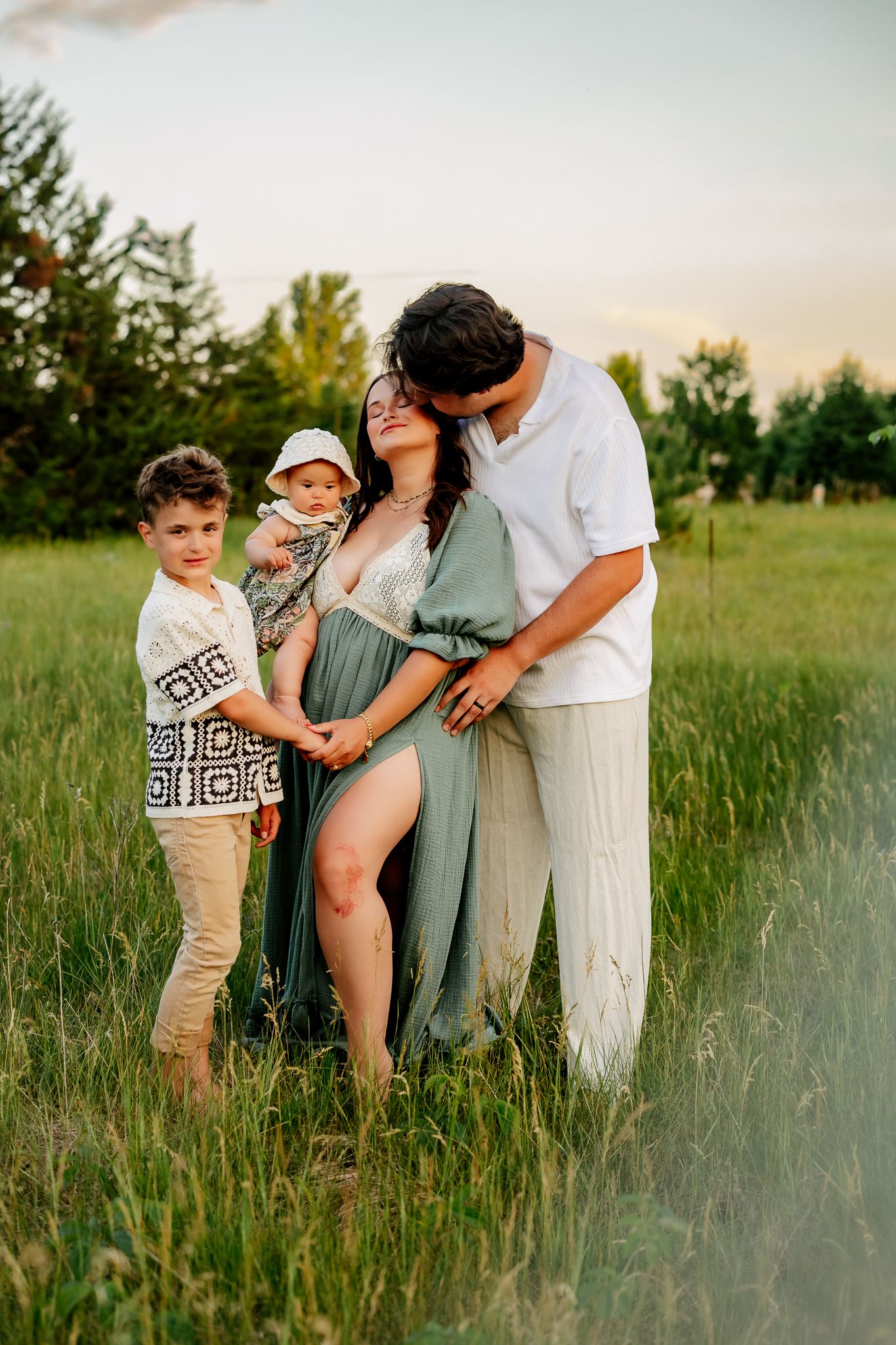 Family maternity portrait at golden hour with pregnant woman in sage green dress embraced by husband and two children.