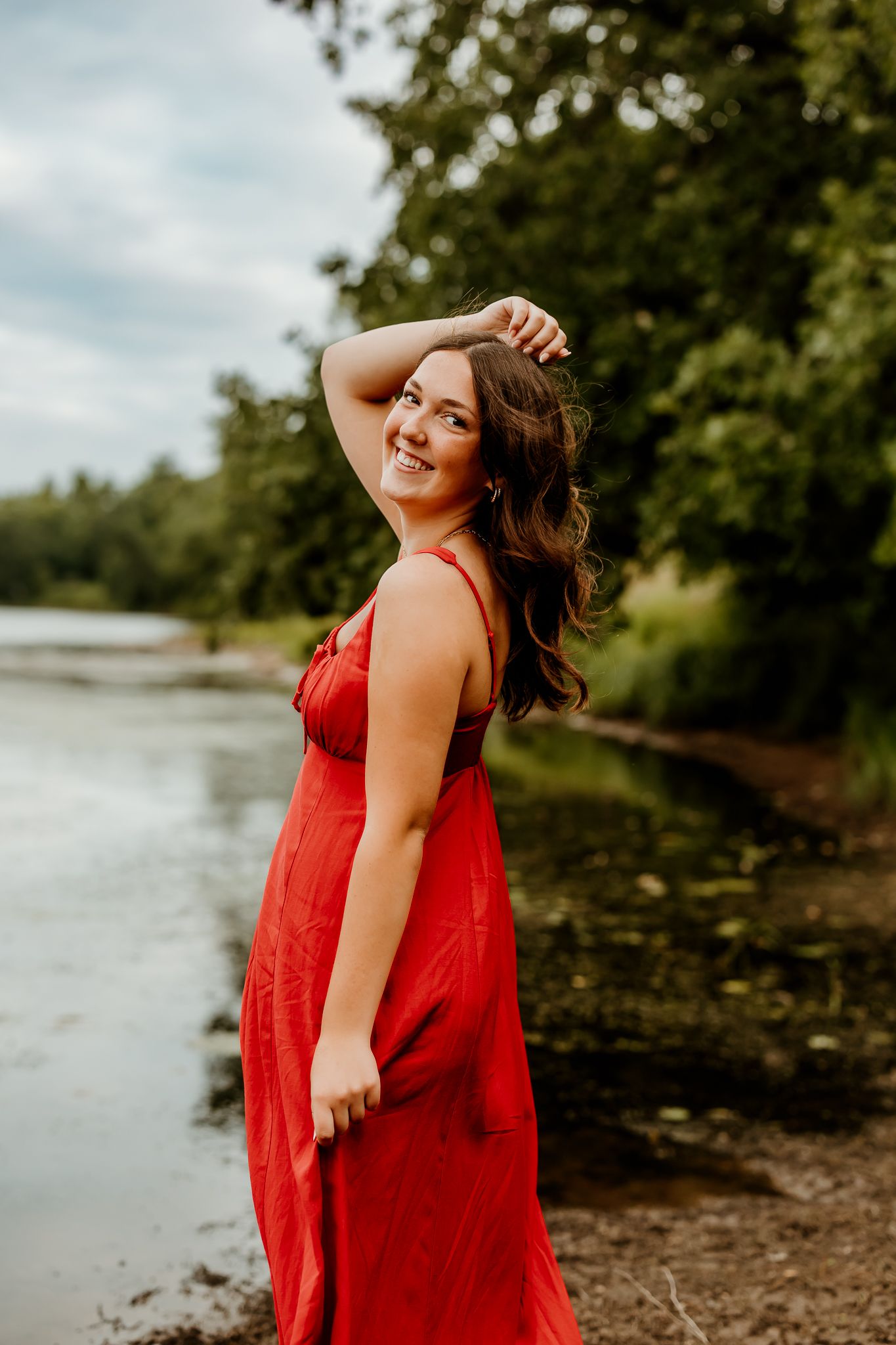 Smiling young woman in a red dress poses by a river surrounded by lush green trees.