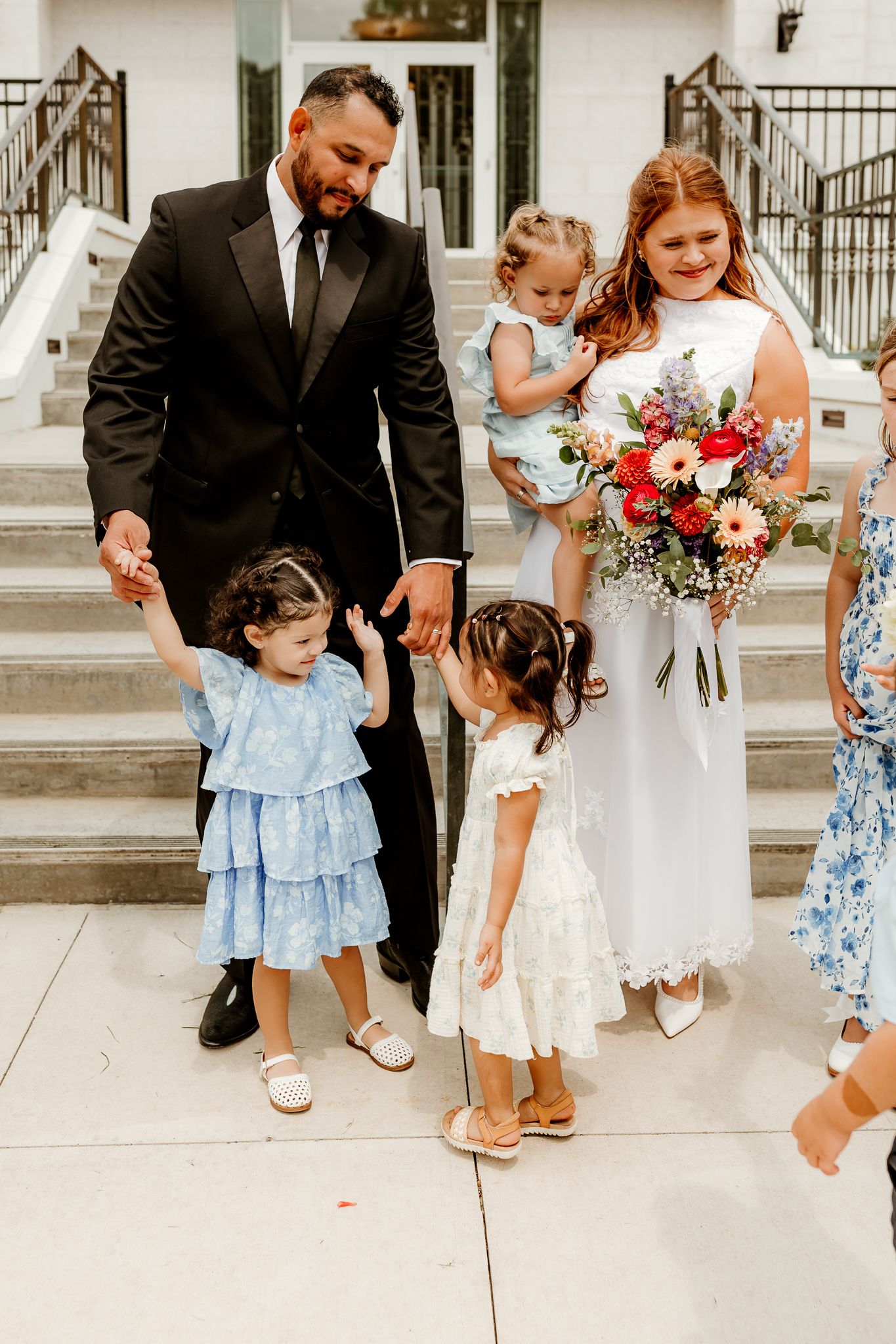 Bride and groom with three young girls in blue and white dresses on wedding day outside white building.