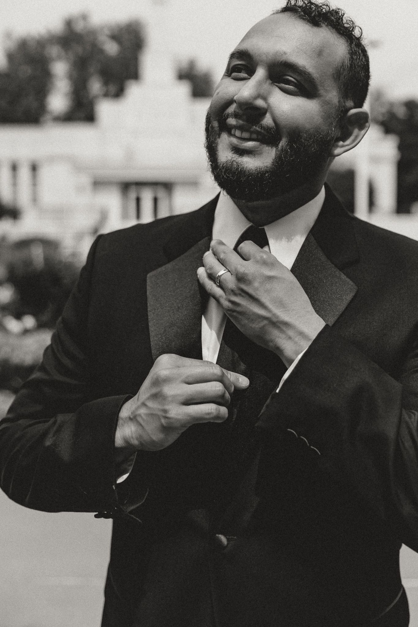 Smiling bearded man in black tuxedo adjusting his tie outdoors in a black and white portrait photo.