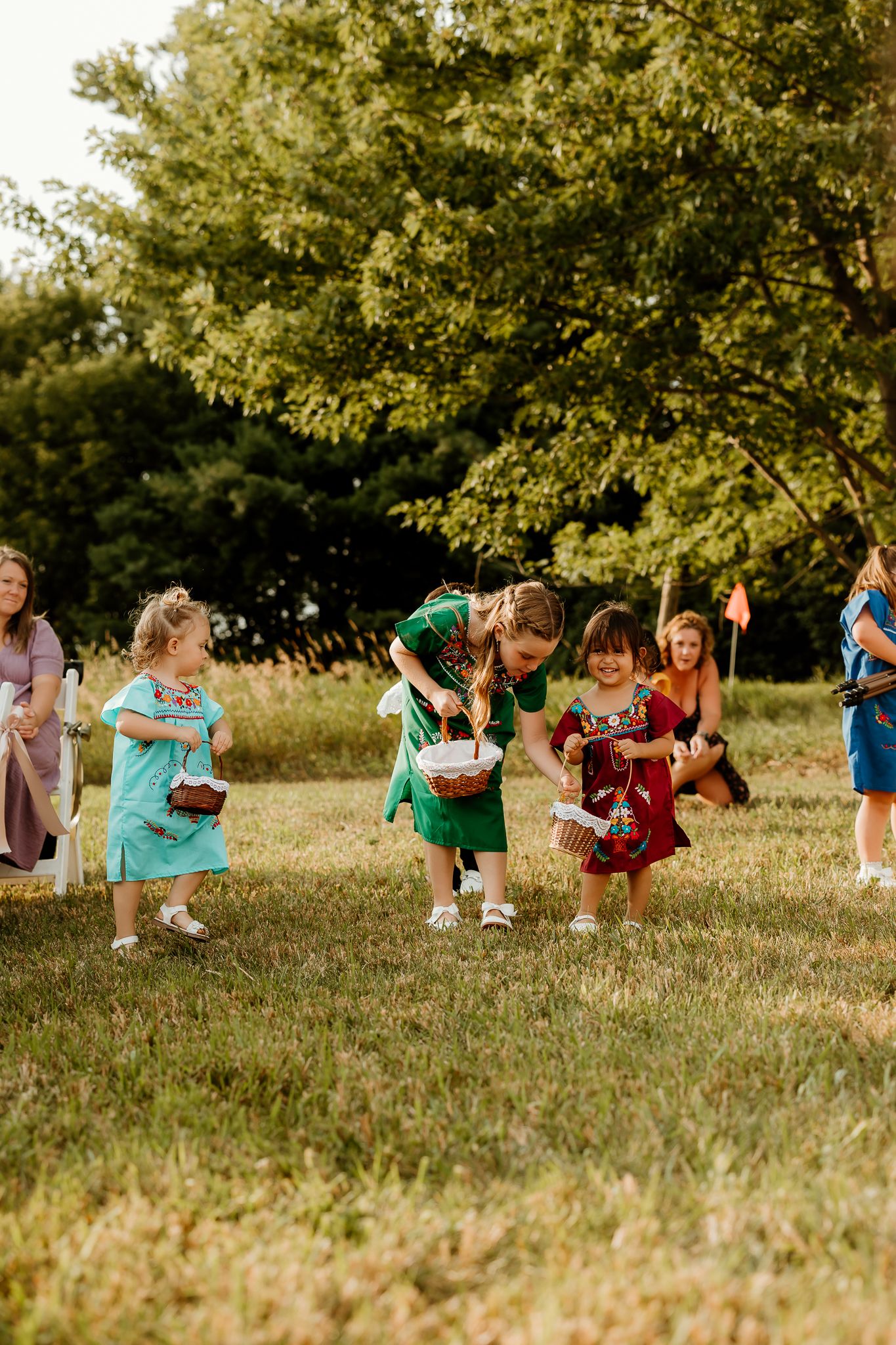 Children in colorful dresses playing outdoors on green grass with trees in the background at a summer gathering.