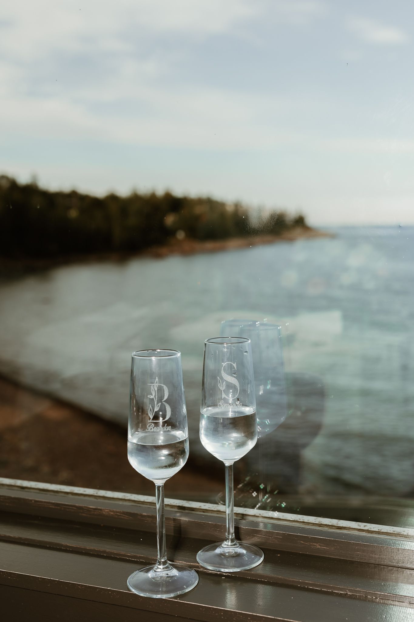 Two monogrammed champagne flutes with water sit on a windowsill overlooking a serene lakeside view.
