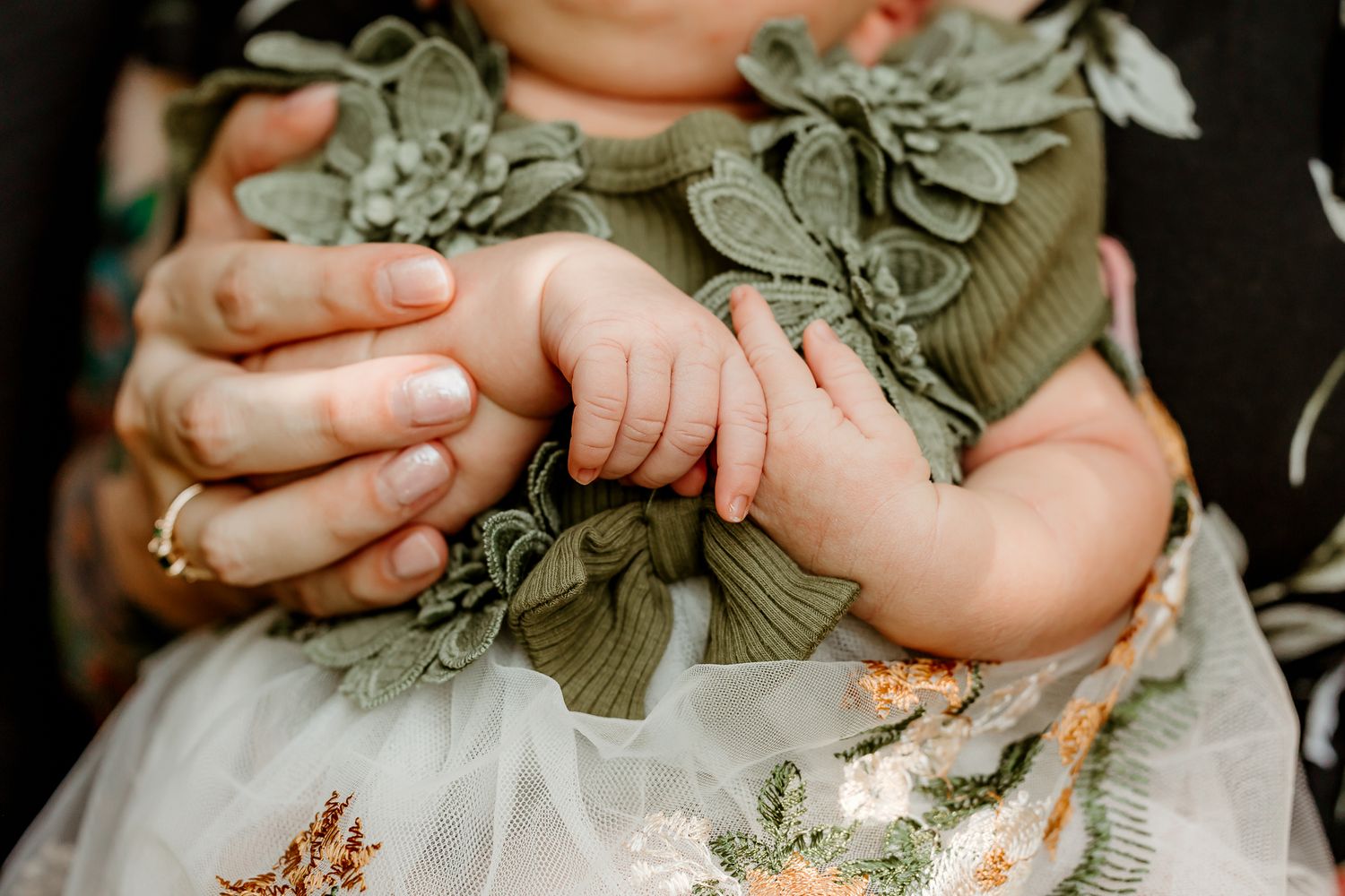 Adult hands gently holding a baby's tiny hands, with the infant wearing a sage green floral dress.