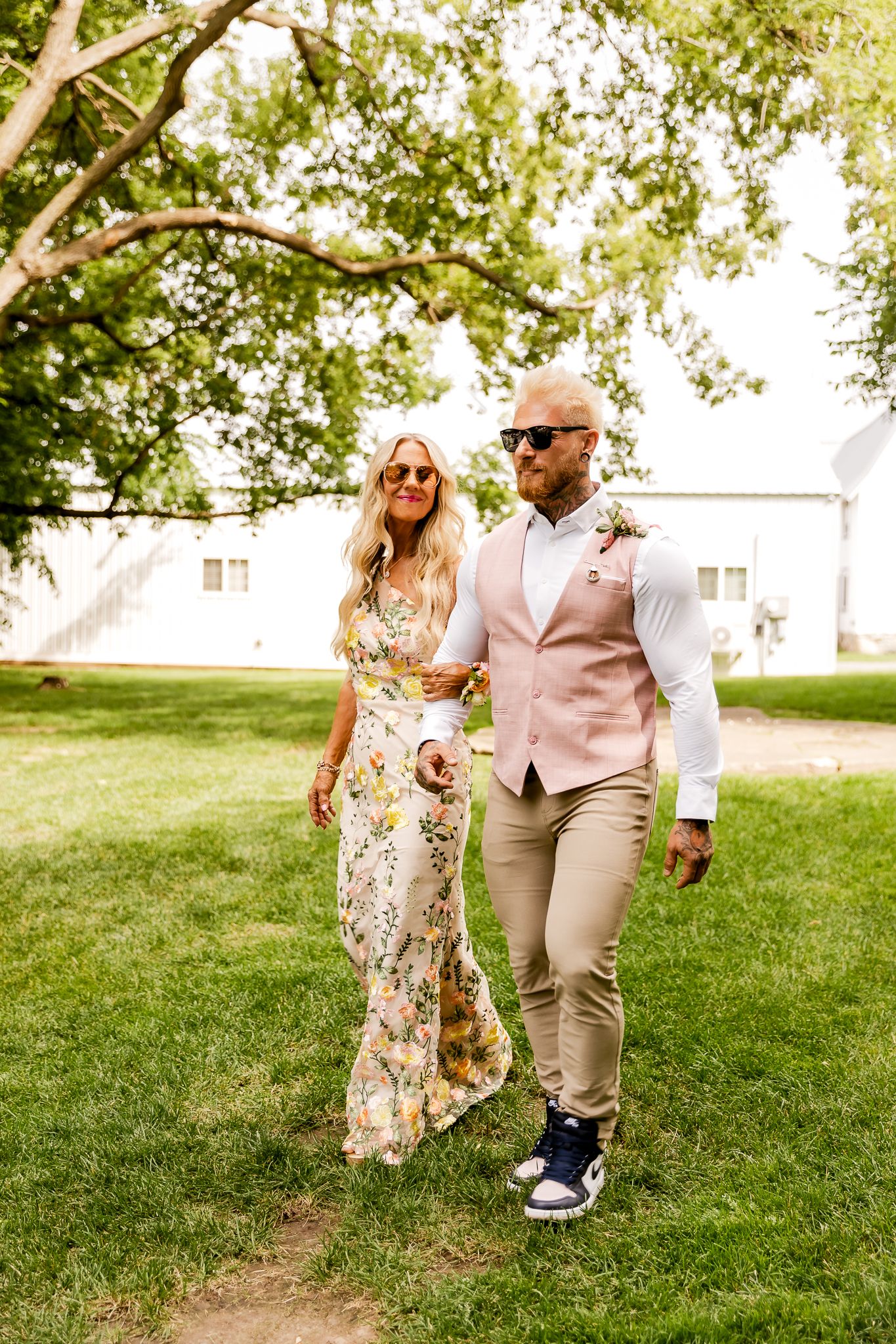 Couple walking outdoors, woman in floral dress, man in pink vest and khakis, both wearing sunglasses, lush green trees behind.