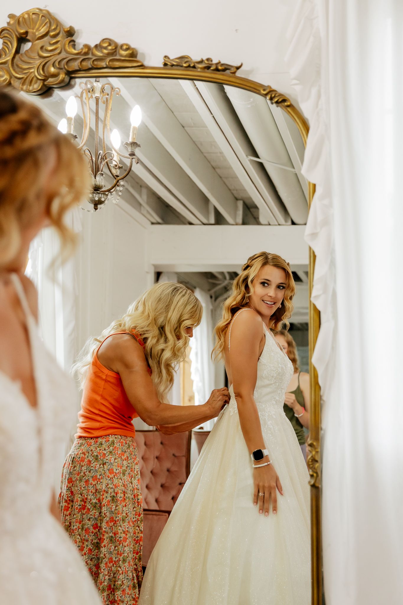 Bride smiling in mirror as woman in orange top helps fasten her white wedding gown in a bright bridal suite.