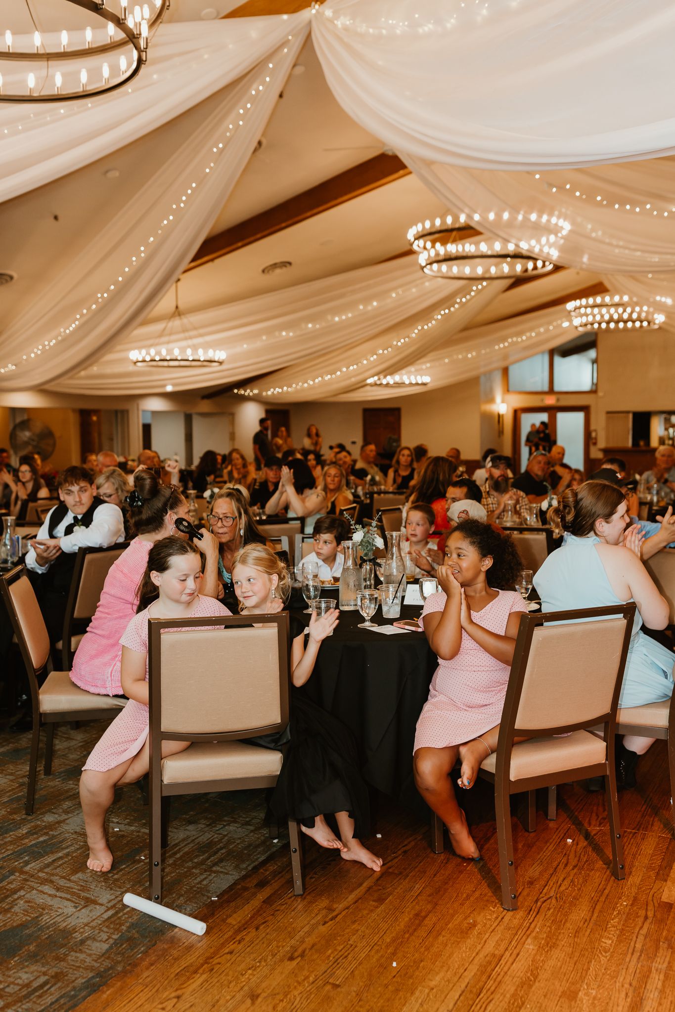 Guests seated at tables in an elegant wedding reception hall with draped fabric ceiling and string lights.