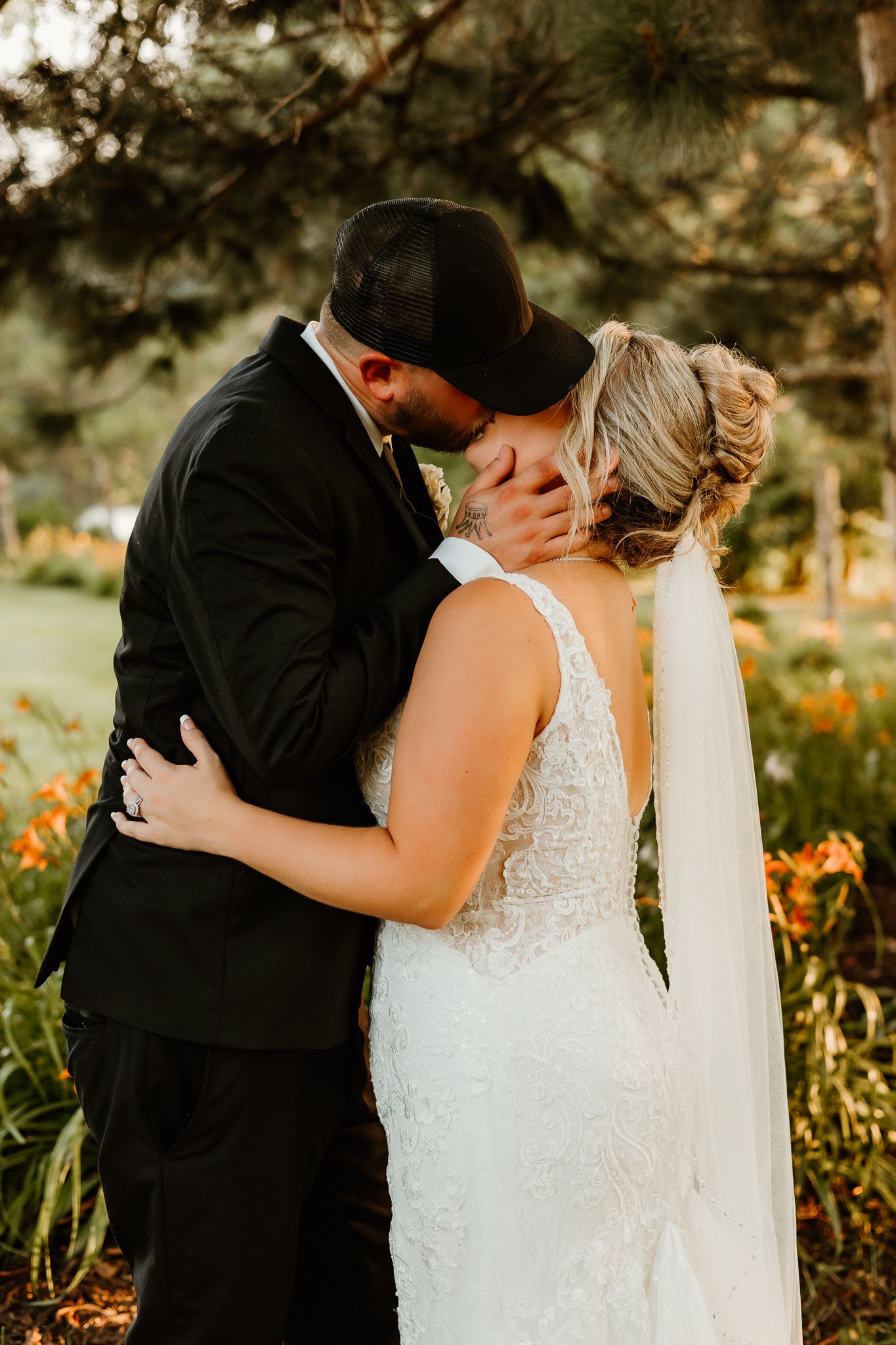 A bride and groom share a romantic kiss outdoors, surrounded by lush greenery and orange flowers during their wedding day.
