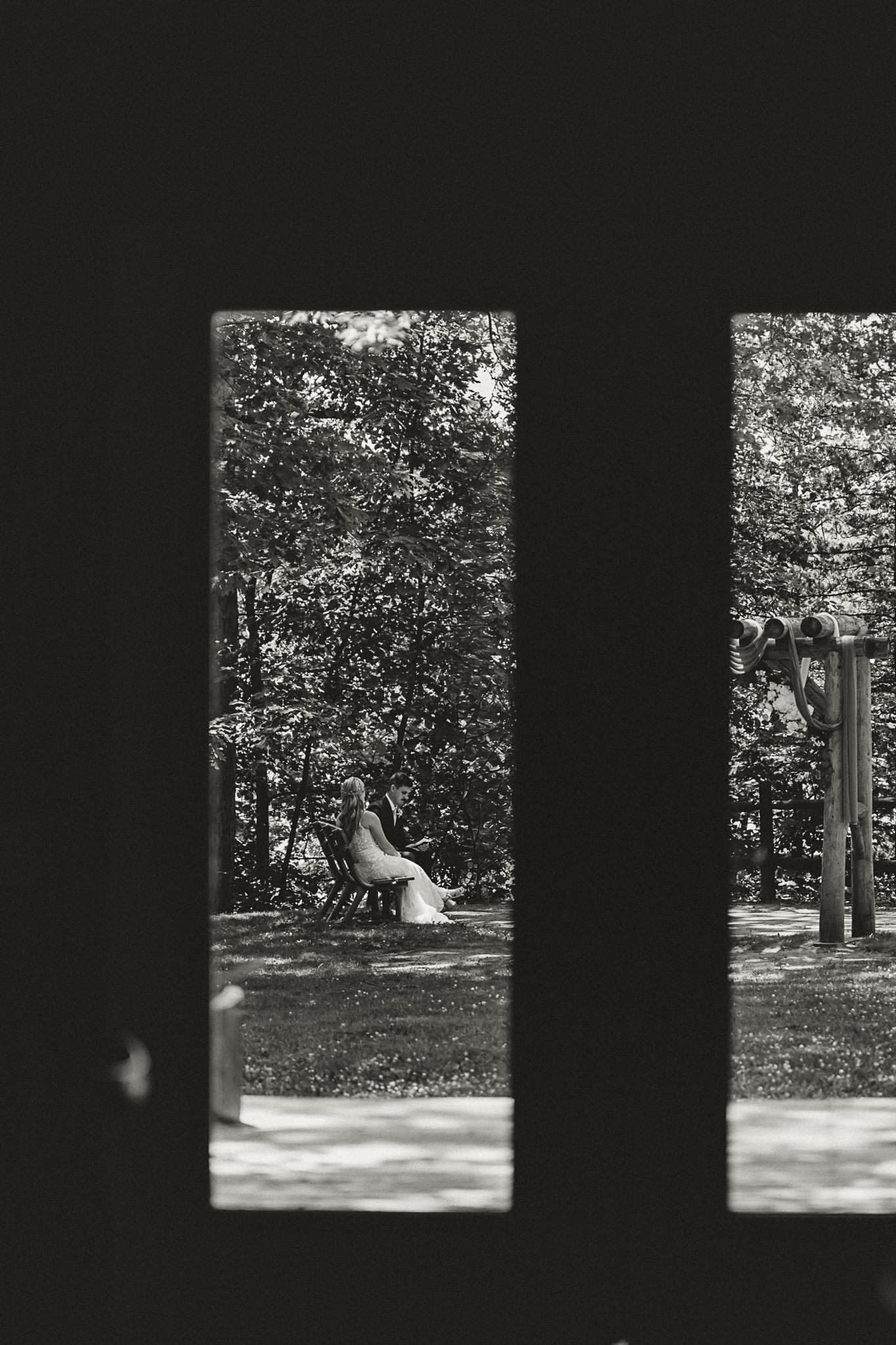 Black and white photo of a bride sitting on a bench in a park, viewed through dark wooden fence gaps.