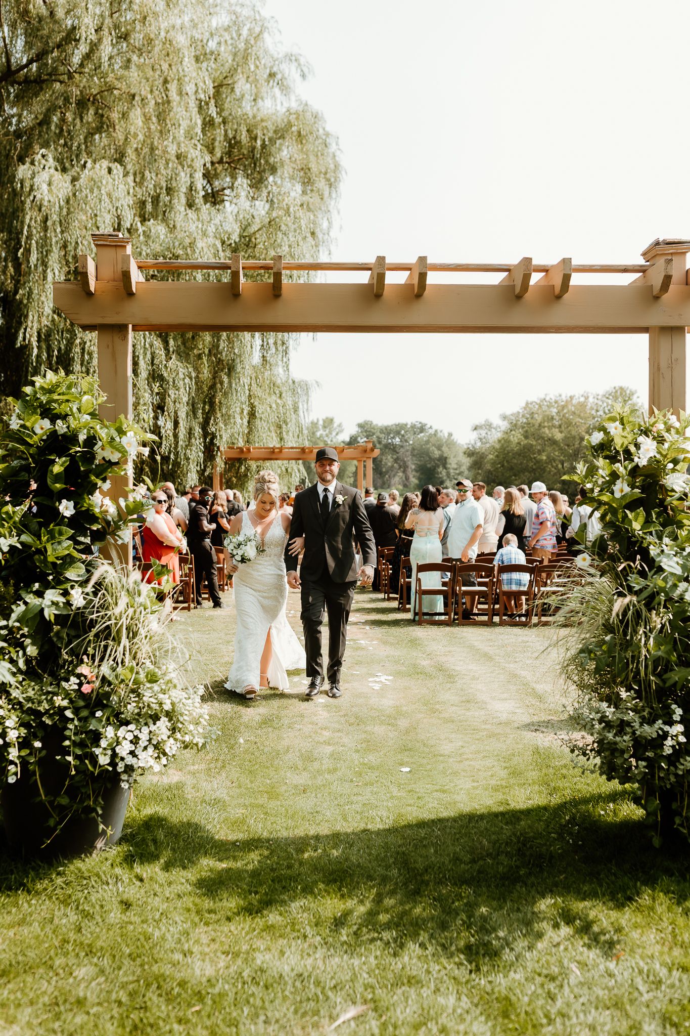 Bride and groom walk down outdoor wedding aisle lined with lush greenery under a wooden pergola on a sunny day.
