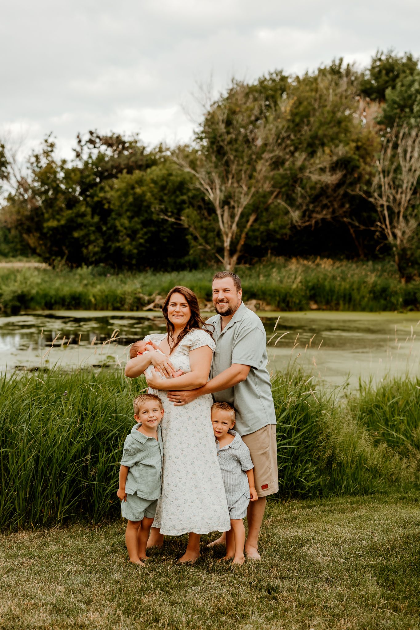 Family of five posing by a pond, parents holding newborn with two young boys in matching sage green outfits.