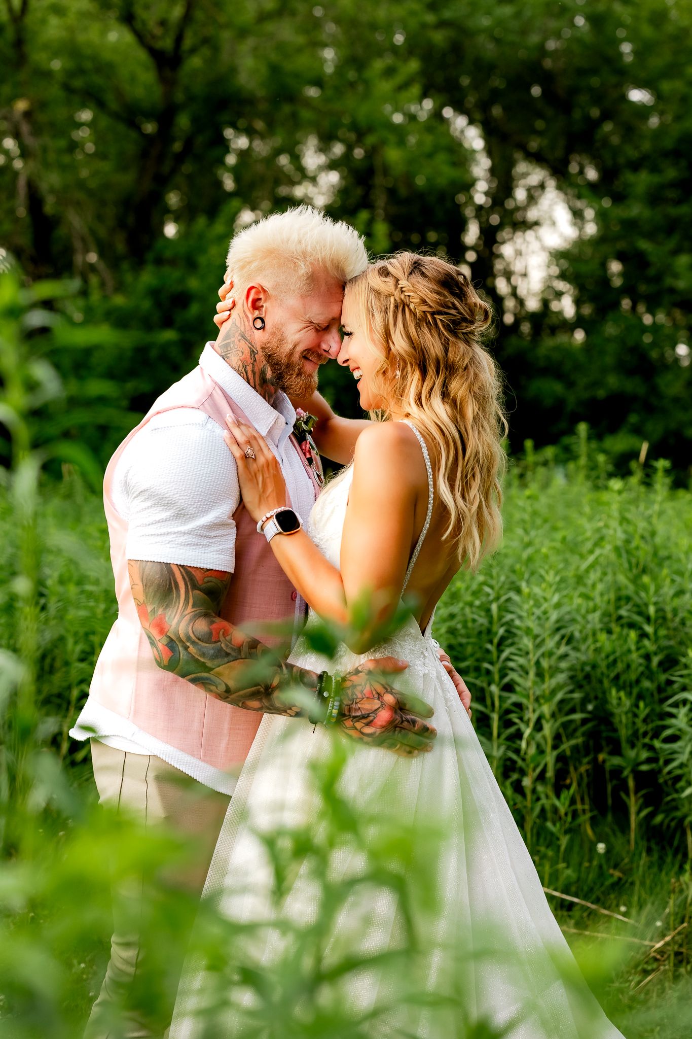 A tattooed groom with blonde hair and a bride in a white dress share a romantic moment outdoors amid lush green foliage.